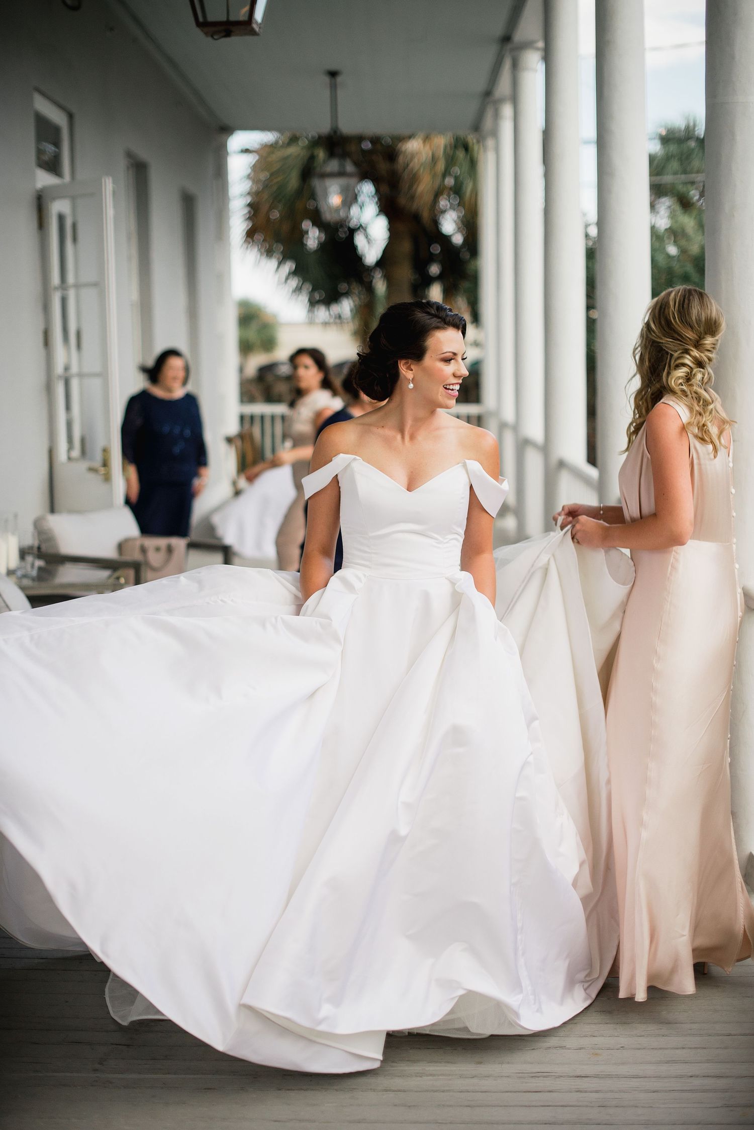Elegant off-shoulder white wedding gown swirls gracefully on a covered porch with bridesmaids in the background.
