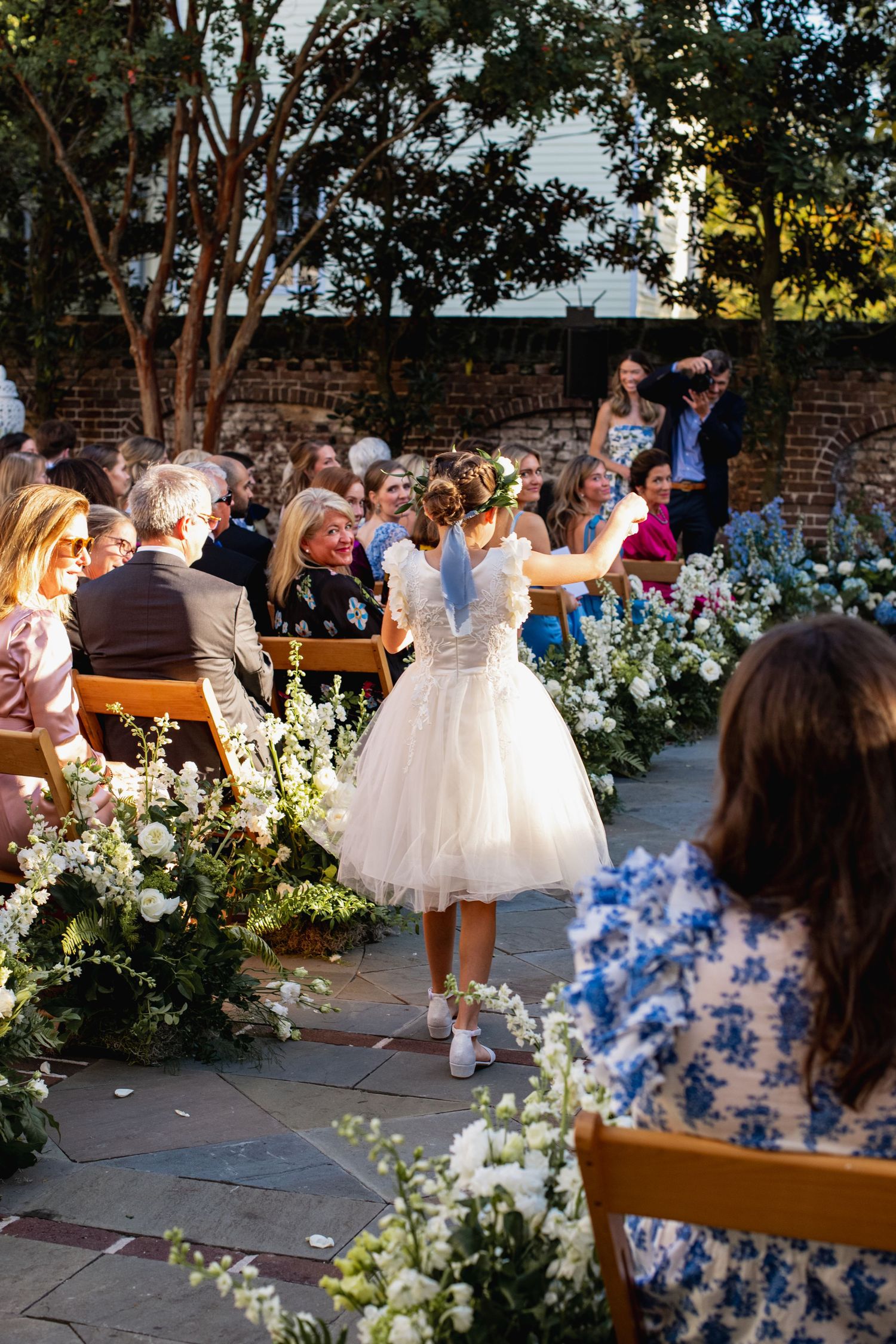 A bride in a tea-length dress and sneakers dances at an outdoor garden wedding ceremony surrounded by white floral arrangements.