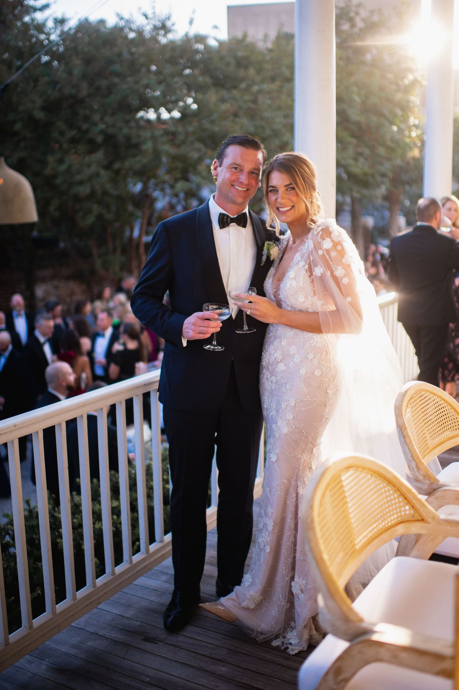 Newlyweds share a romantic moment on a white porch with sunlight streaming through the trees at sunset.