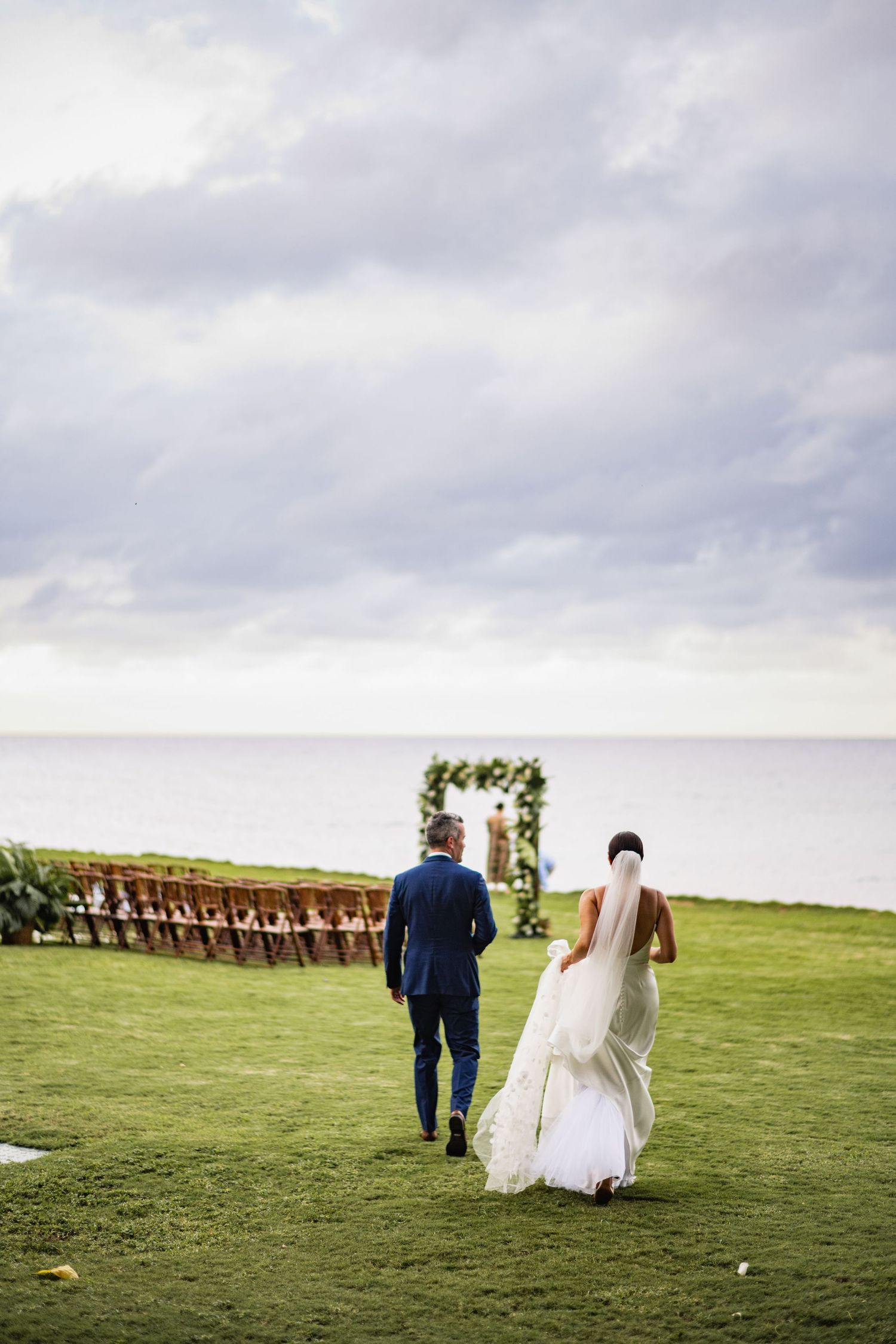 A couple walks across a green hillside field under cloudy skies during their wedding celebration.