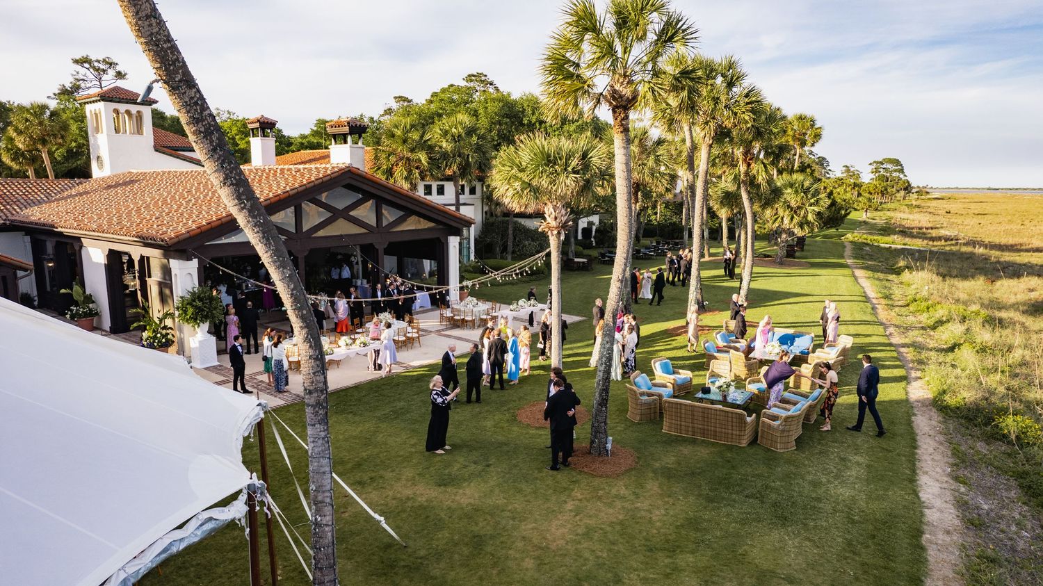 A panoramic view of a rustic barn venue with palm trees and guests gathered for an outdoor celebration.