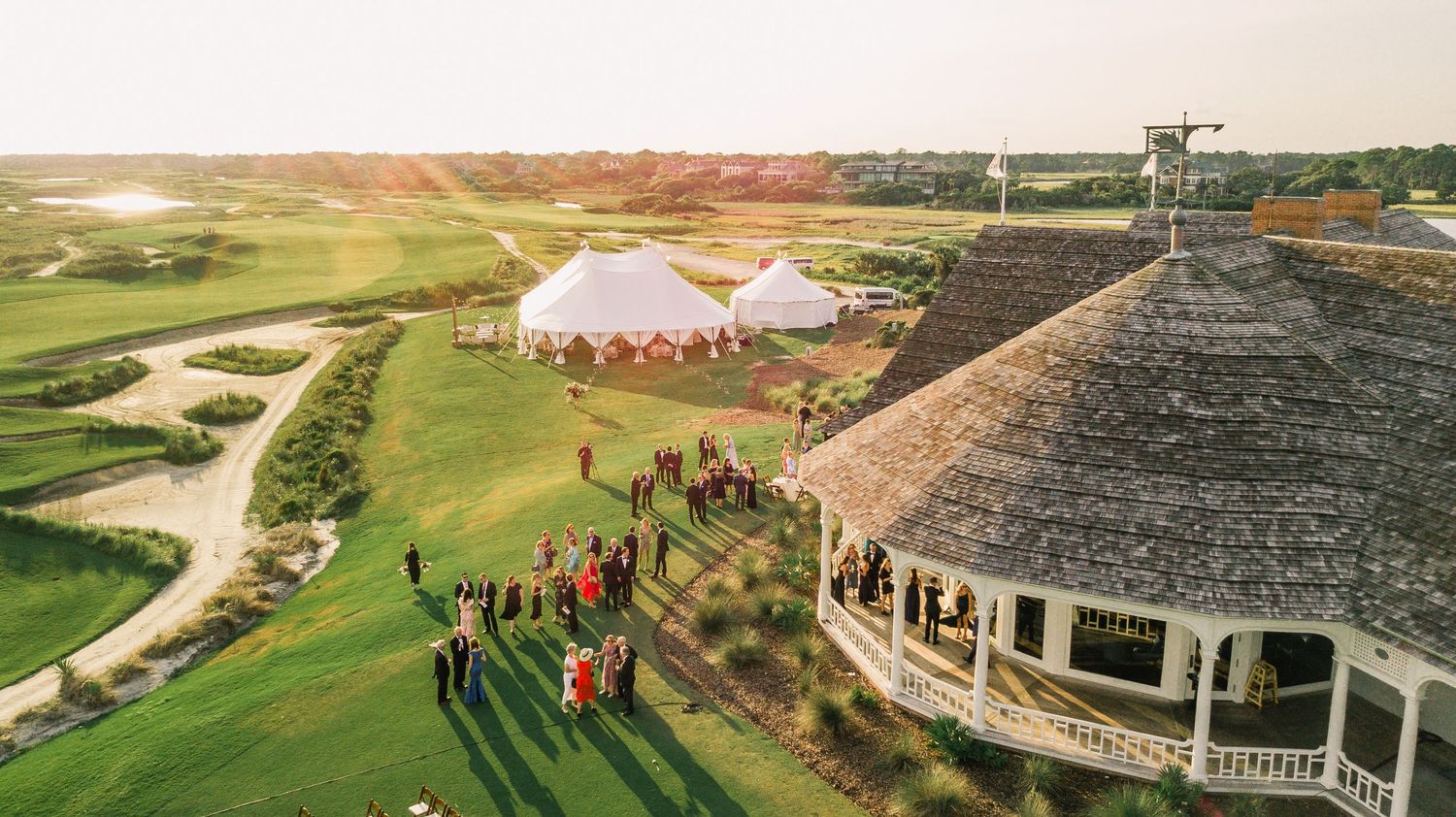 Aerial view of a golf course venue with thatched roof gazebo and gathering of people during sunset.