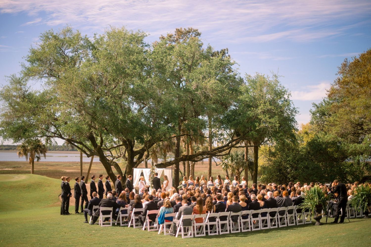 Panoramic view of an outdoor wedding ceremony setup with white chairs on a lush green golf course under blue skies.