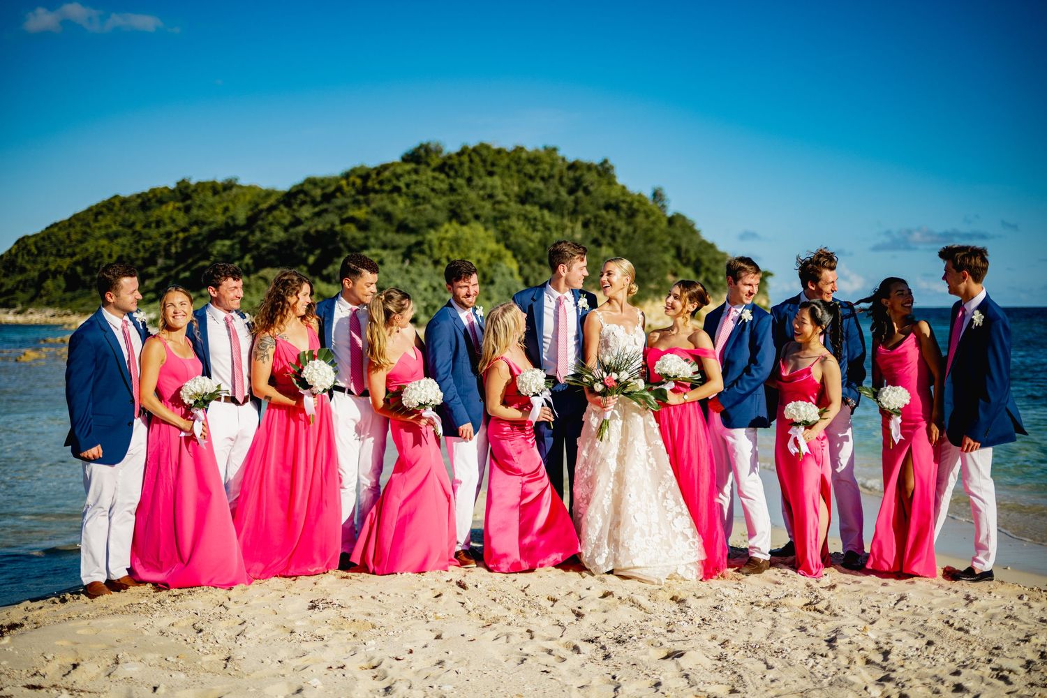 Wedding party in pink bridesmaid dresses and white attire poses together on tropical beach.