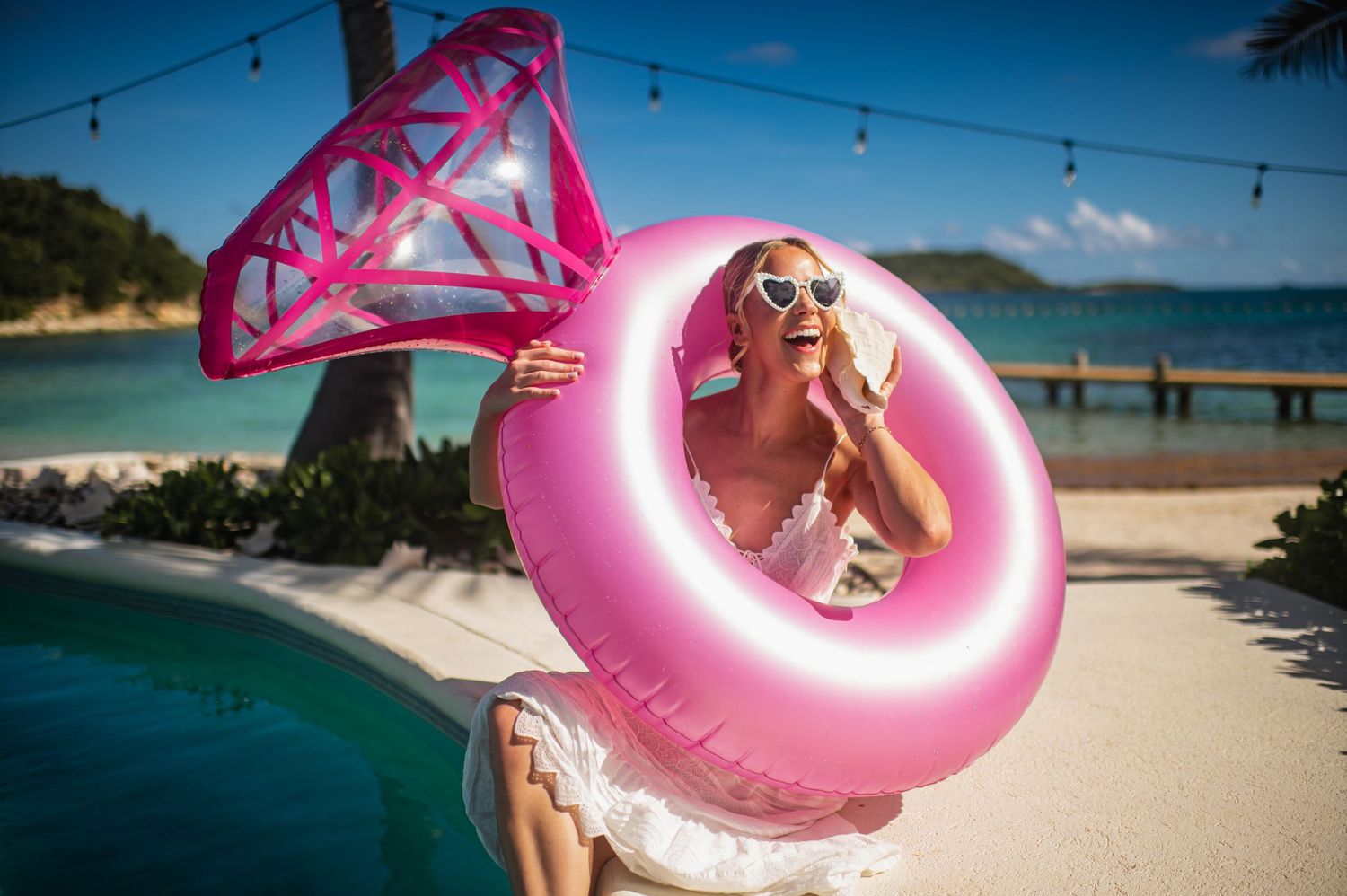 Someone holds a bright pink inflatable pool float against a sunny beach backdrop with turquoise water and sand.
