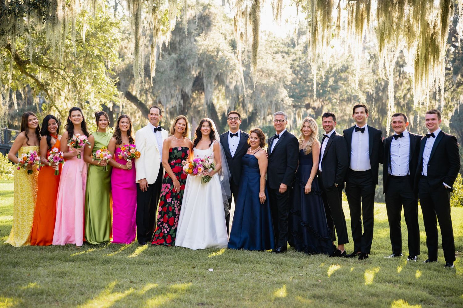 Large wedding party poses together outdoors under Spanish moss draped trees in a garden setting.