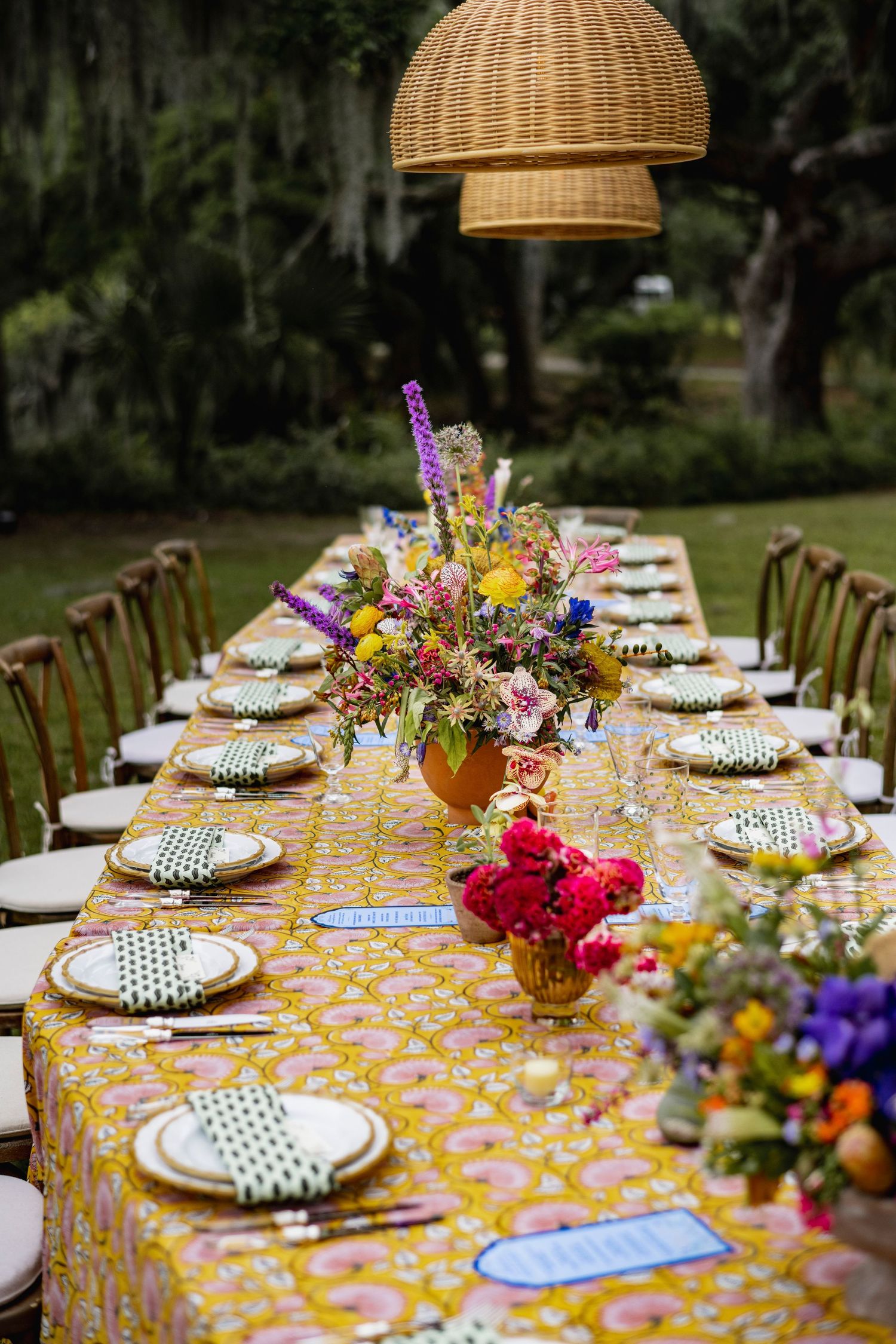 Elegant outdoor table setting with wicker pendant lamps, polka dot plates and colorful floral centerpieces on yellow tablecloth.