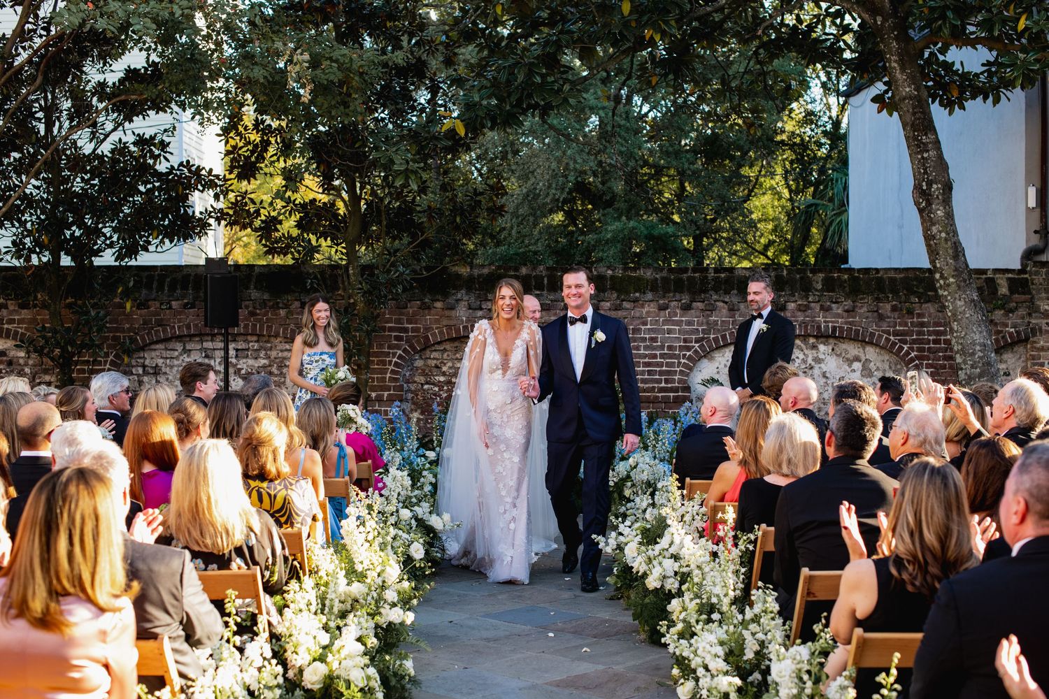 An outdoor wedding ceremony with guests seated on wooden chairs along a flower-lined aisle against a rustic brick wall.