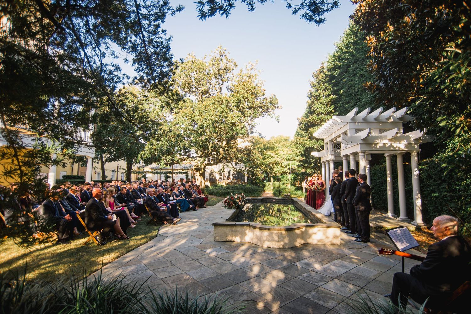 Panoramic view of an outdoor wedding ceremony under a white pergola with guests seated on white chairs in a garden setting.
