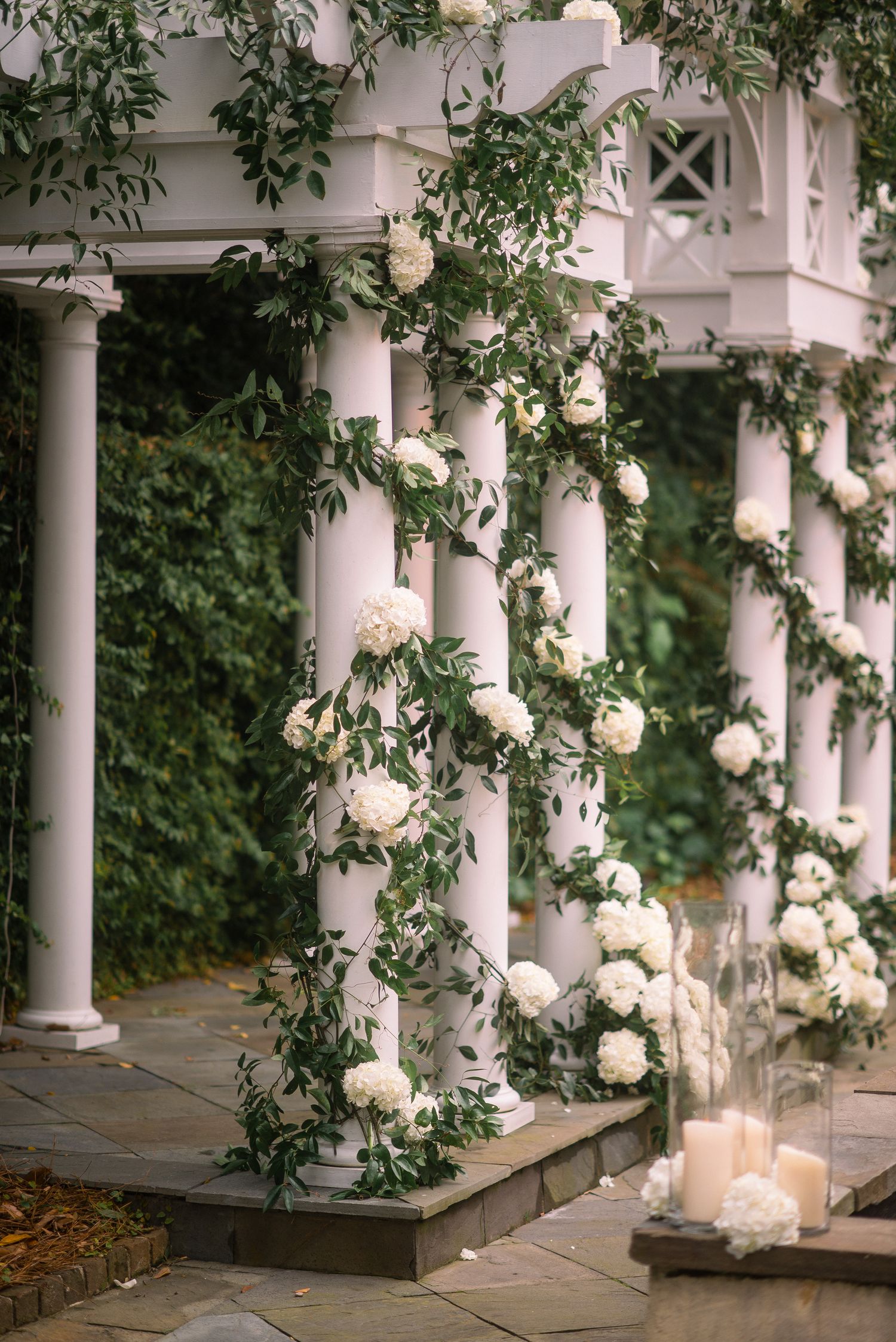 White roses and greenery adorn classic columns along an elegant outdoor patio with glowing candles.