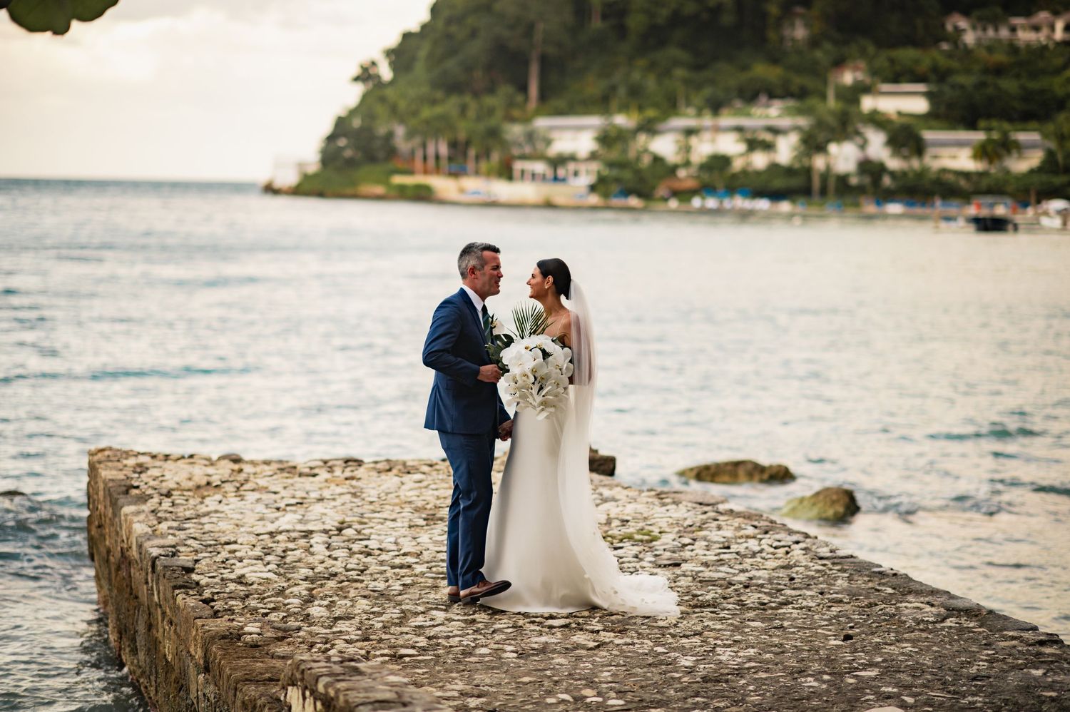 Couple in wedding attire sharing romantic moments along a scenic coastal shoreline at sunset.