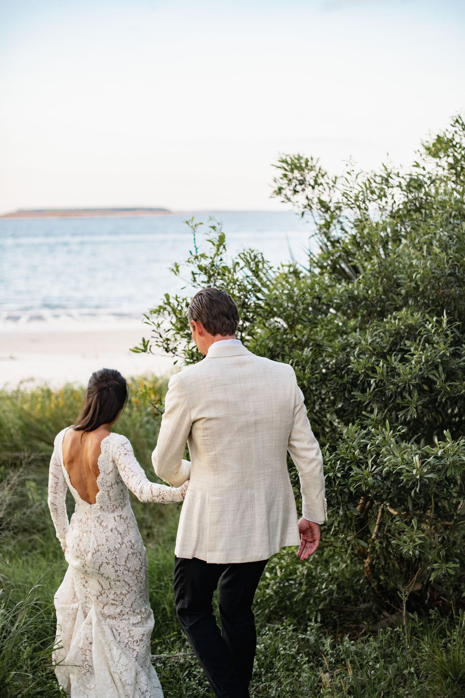 A couple in wedding attire walks together through lush greenery near a beach on their special day.