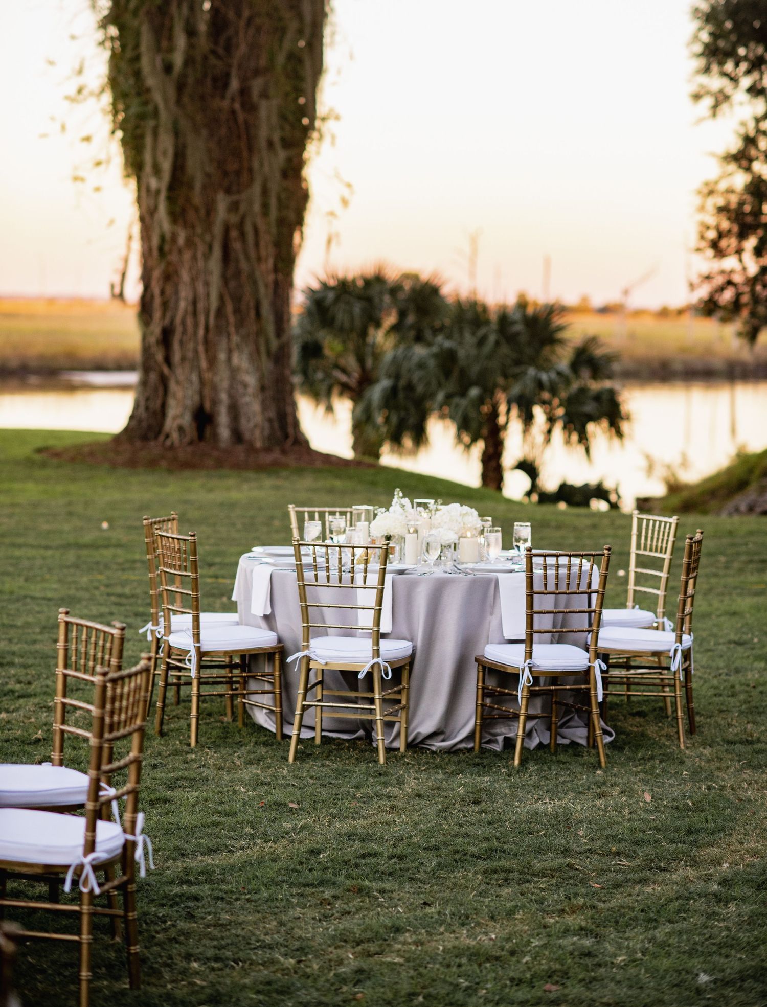 Elegant outdoor wedding reception setup with multiple round tables and gold chiavari chairs on a grassy lawn at sunset.