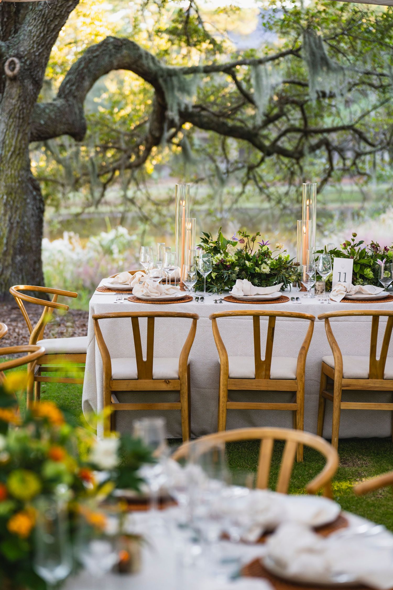 Elegant outdoor wedding reception table setup with wooden chairs and white linens under oak trees at golden hour.