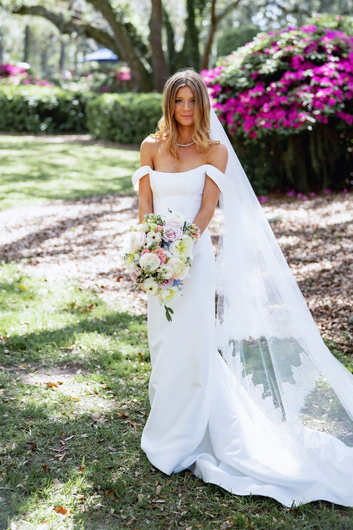 Bride in flowing white dress poses with bouquet among blooming azalea gardens.