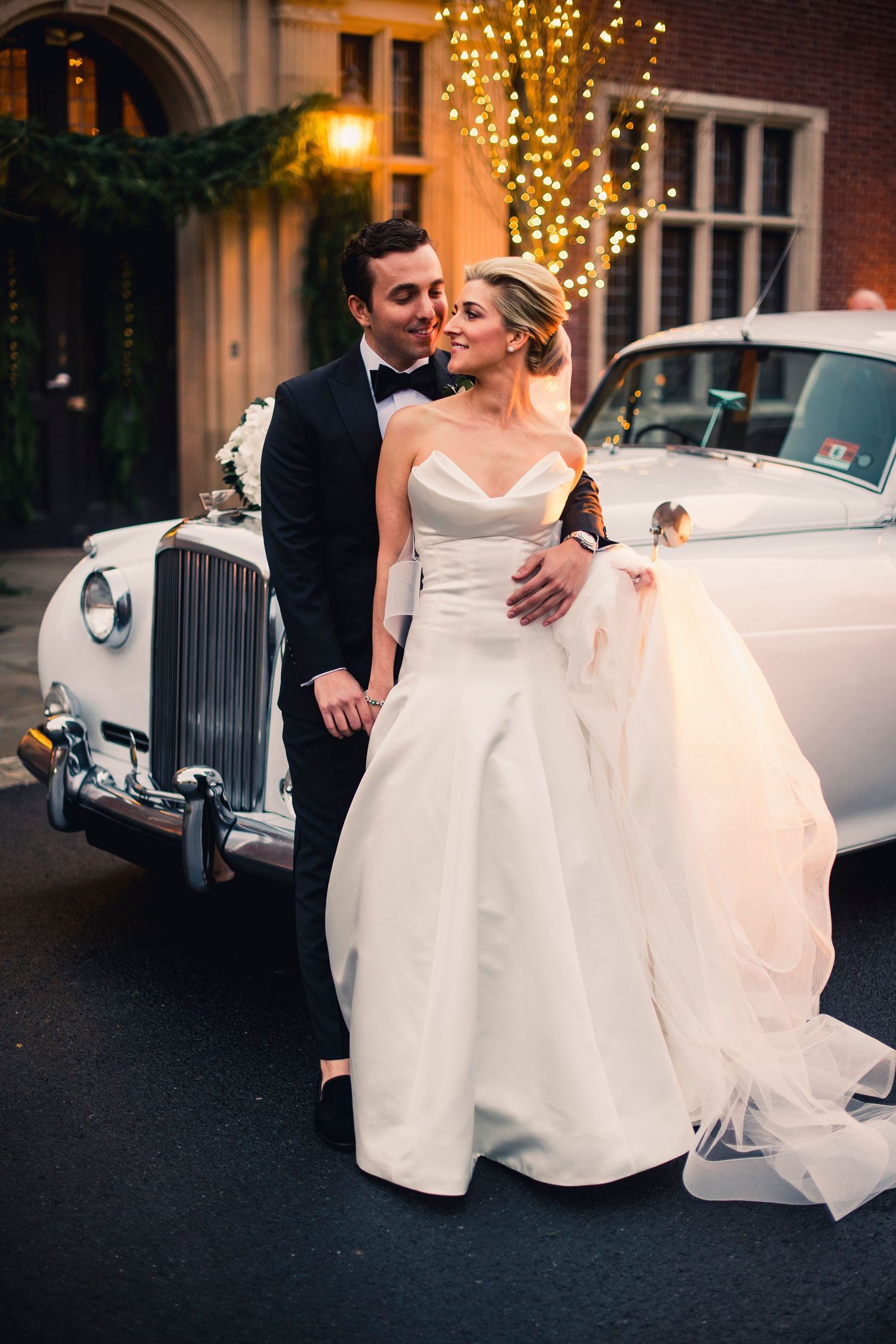 Elegant couple poses by vintage white Rolls Royce under romantic evening lighting.