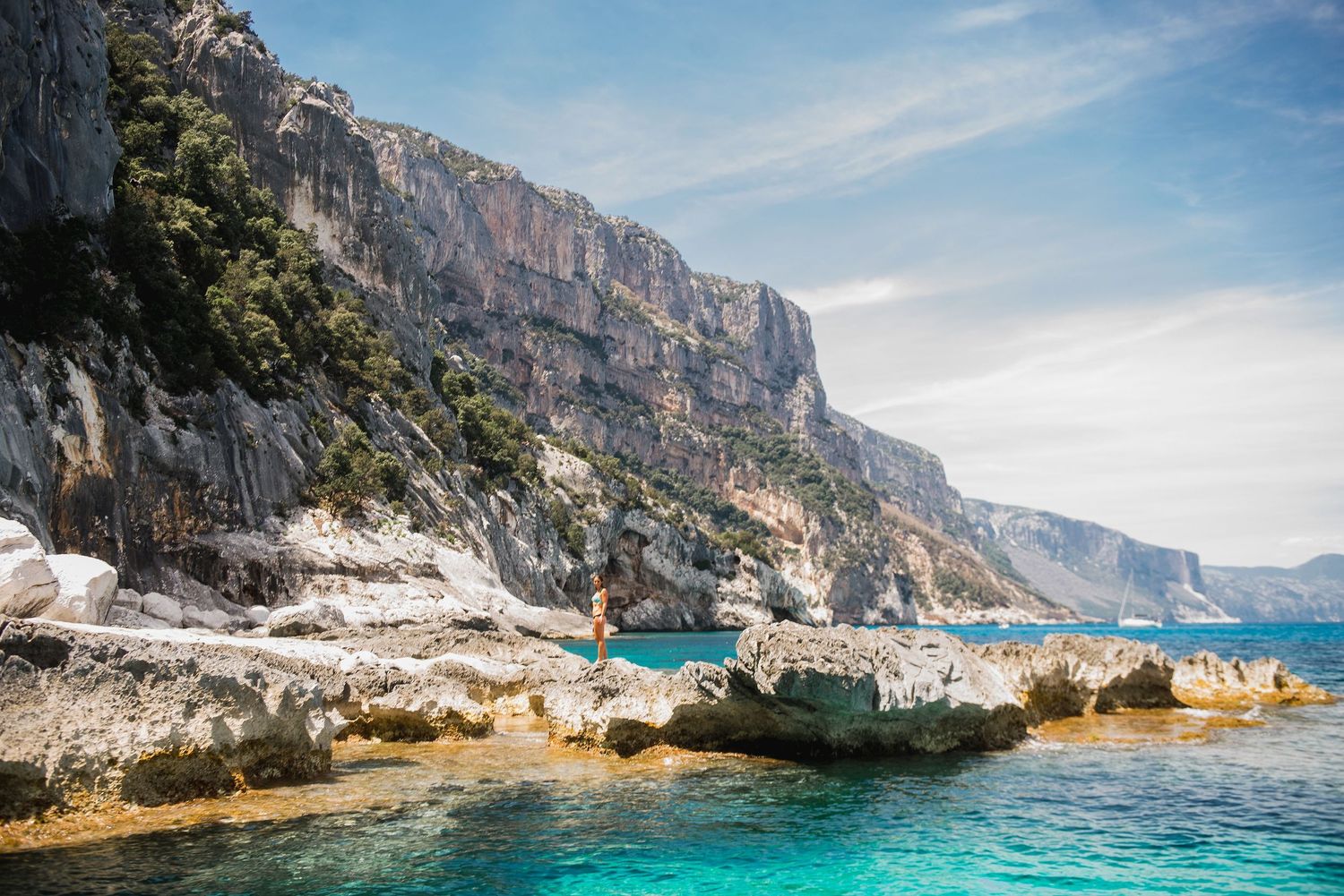 Dramatic limestone cliffs tower over turquoise Mediterranean waters along a rocky coastline.