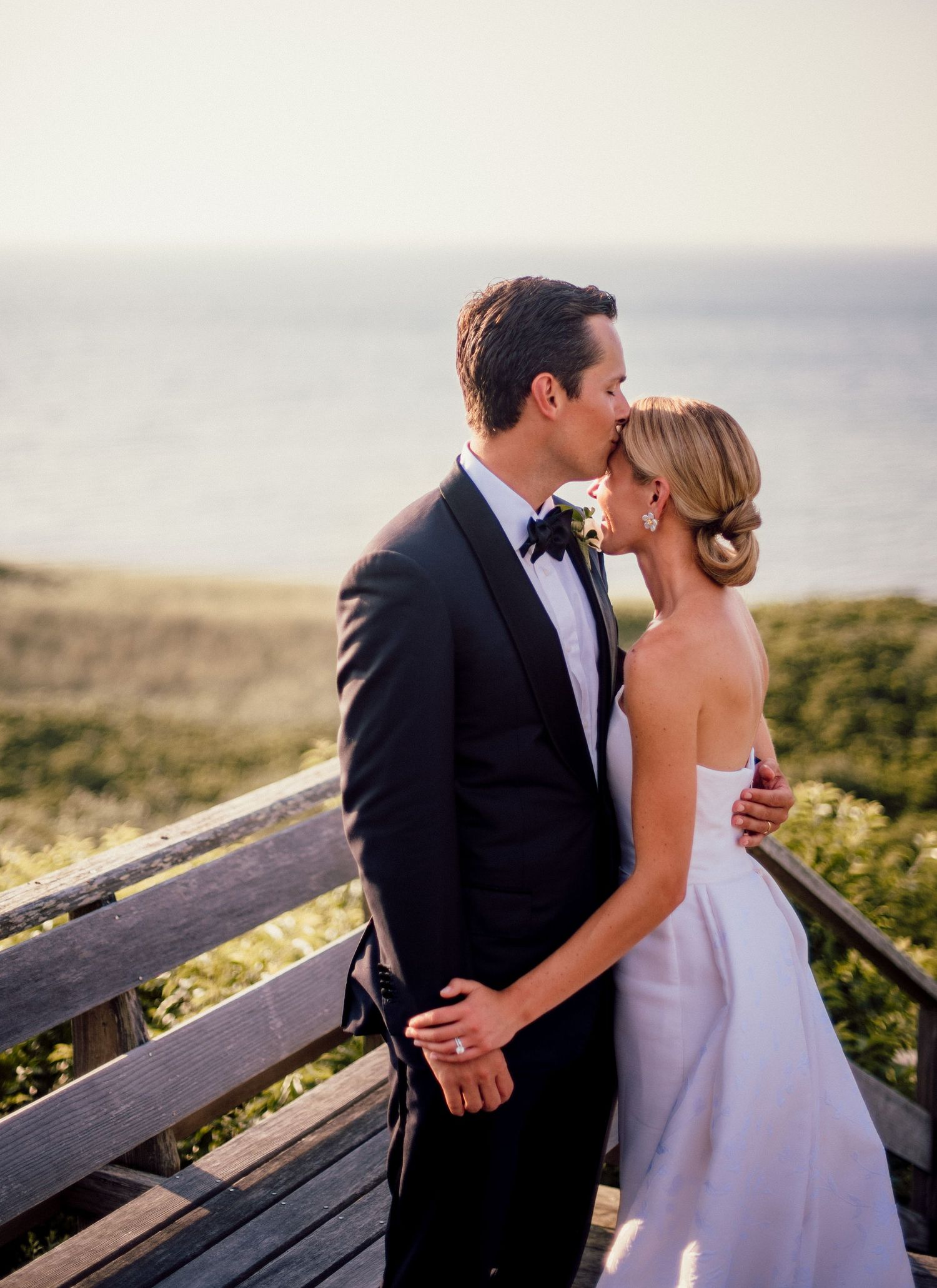 A couple shares a romantic moment on a wooden deck overlooking a scenic coastal landscape at sunset.