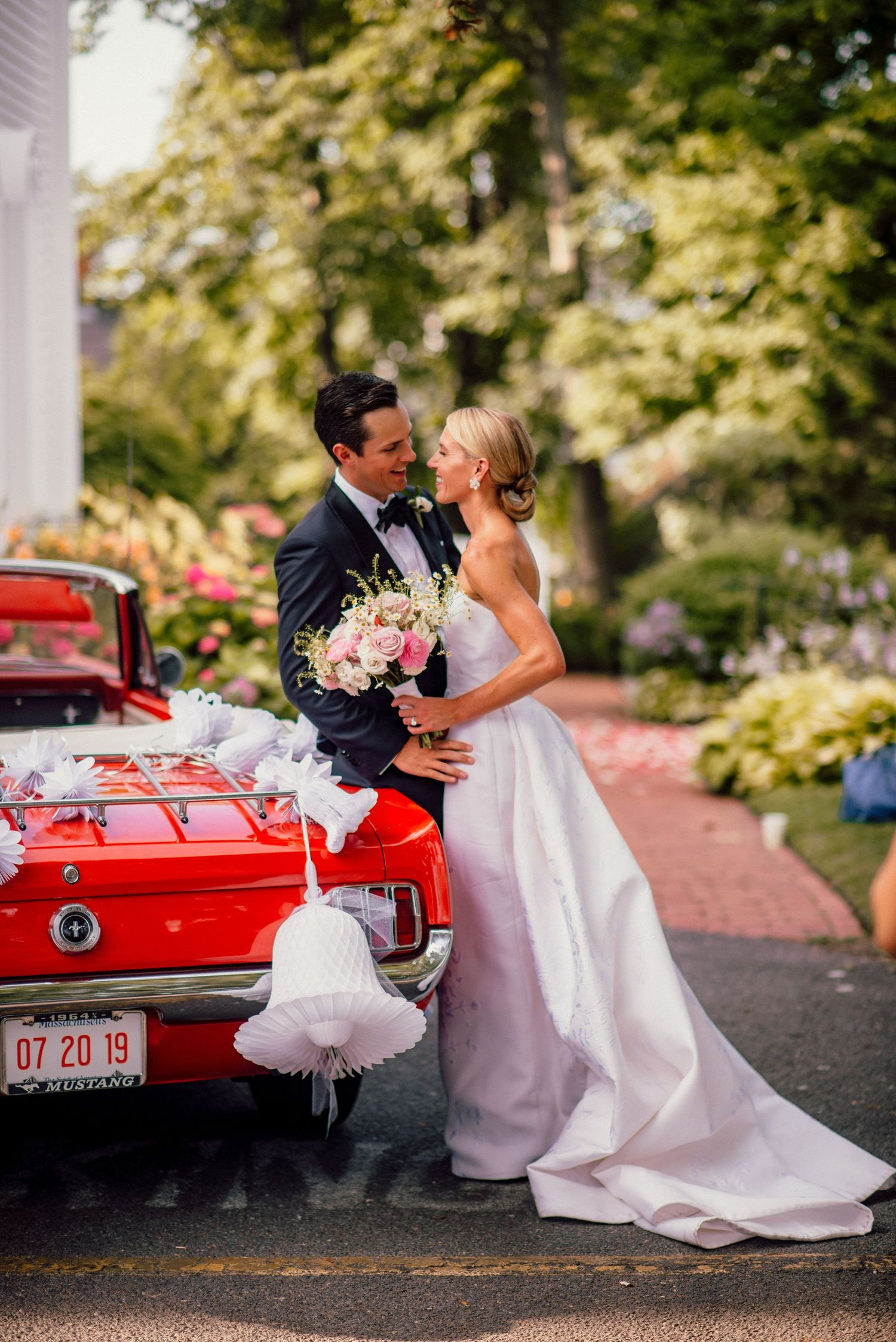 Romantic wedding photo with couple posed against vintage red car surrounded by lush gardens.