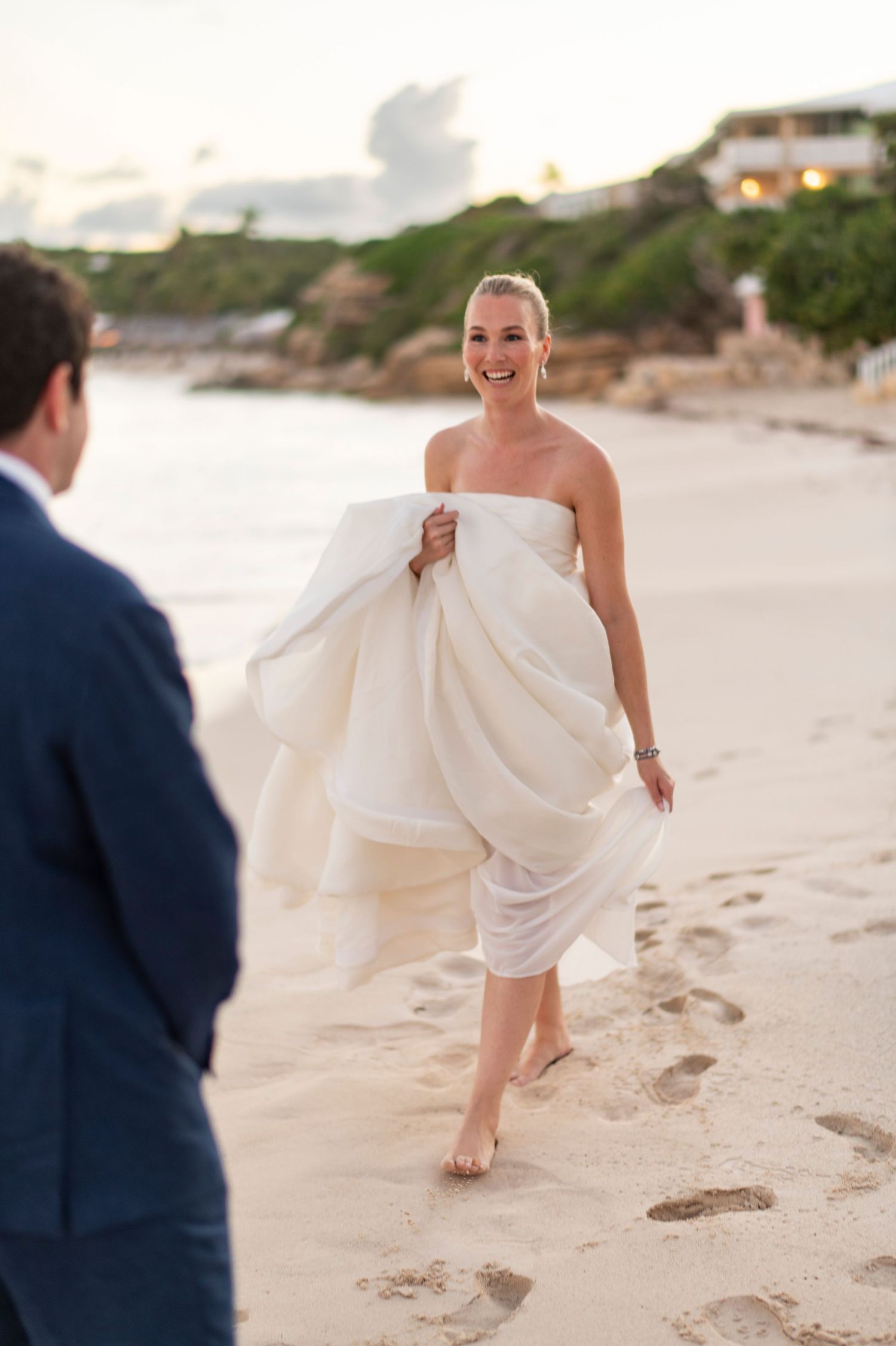 A bride in white dress runs playfully along a sandy beach during wedding photos.