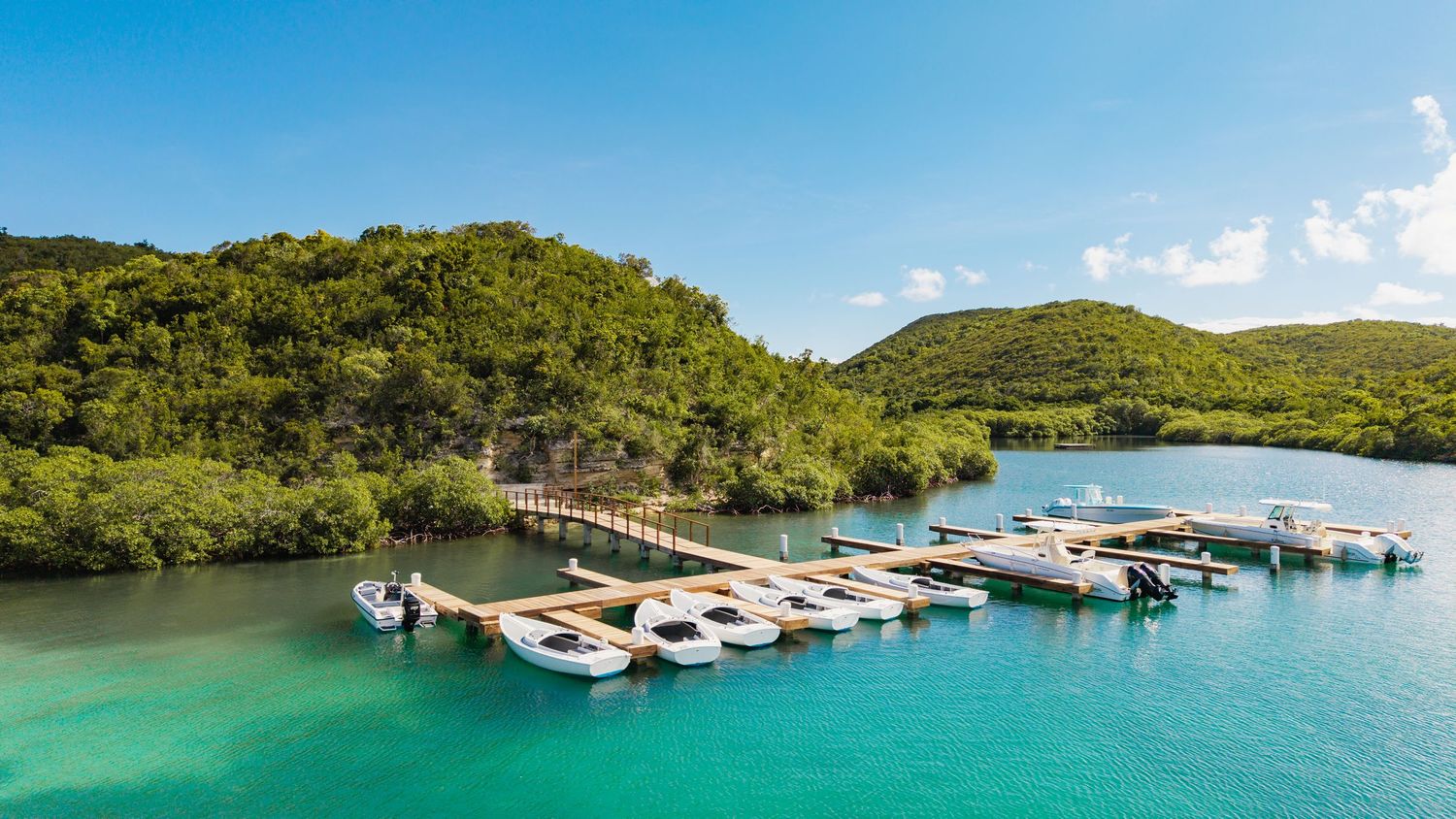 Aerial view of a boat dock with multiple vessels moored in pristine turquoise waters surrounded by green hills.