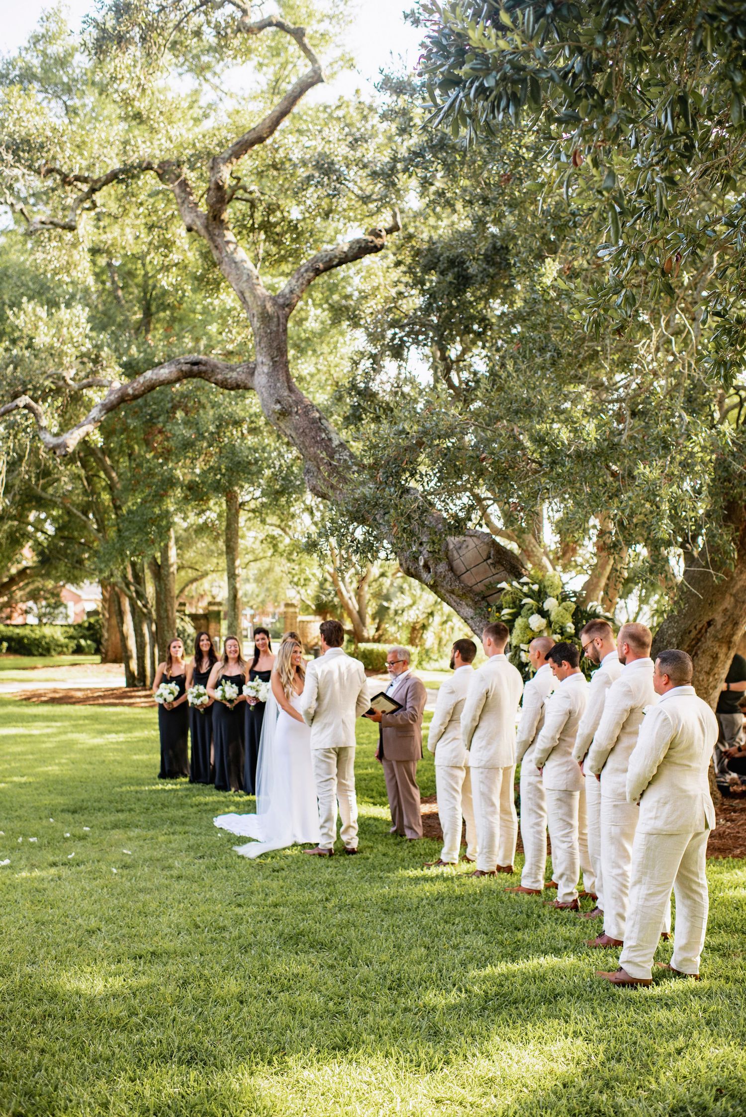 Wedding party members in white attire gather beneath oak trees on a sunlit grassy lawn during an outdoor ceremony.