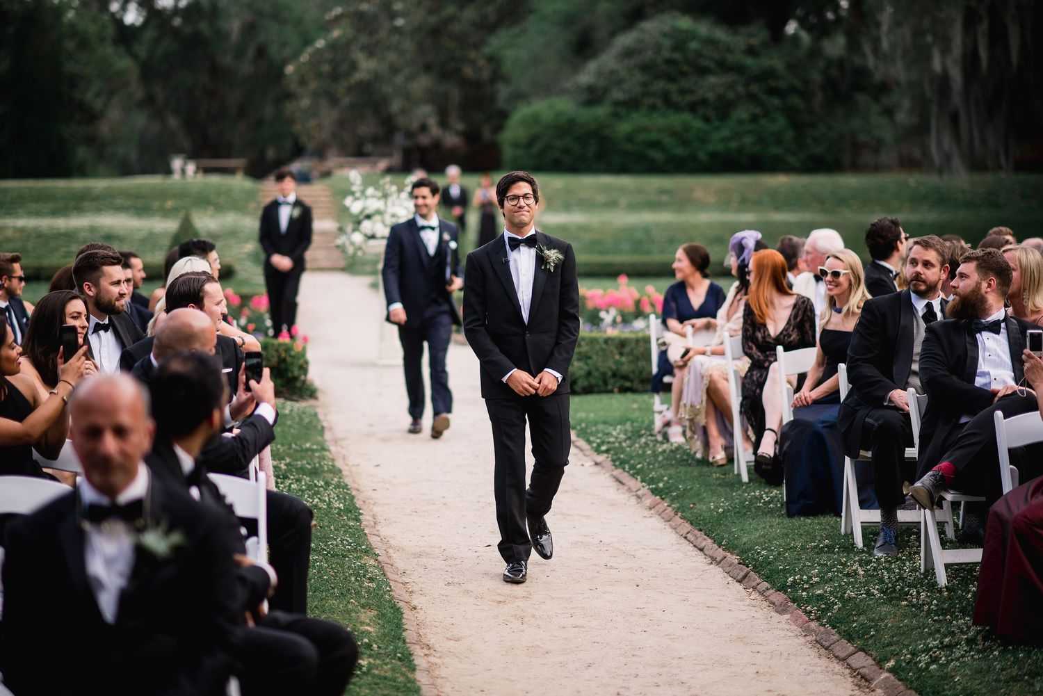 Wedding guests gather outdoors for a garden ceremony with rows of chairs set up on a lush green lawn.
