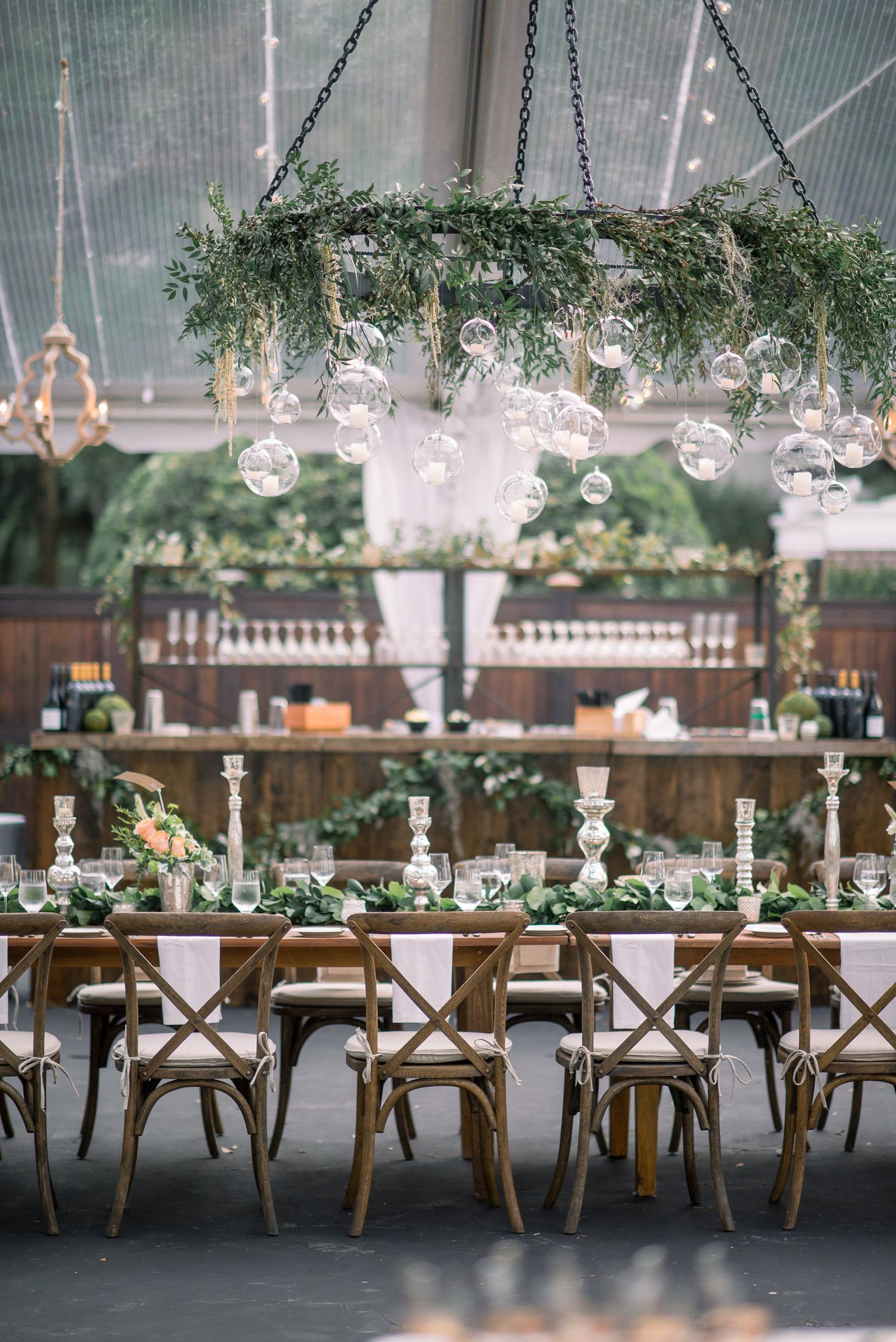 Long rustic wooden dining tables with cross-back chairs set up for an elegant wedding reception under hanging lights.