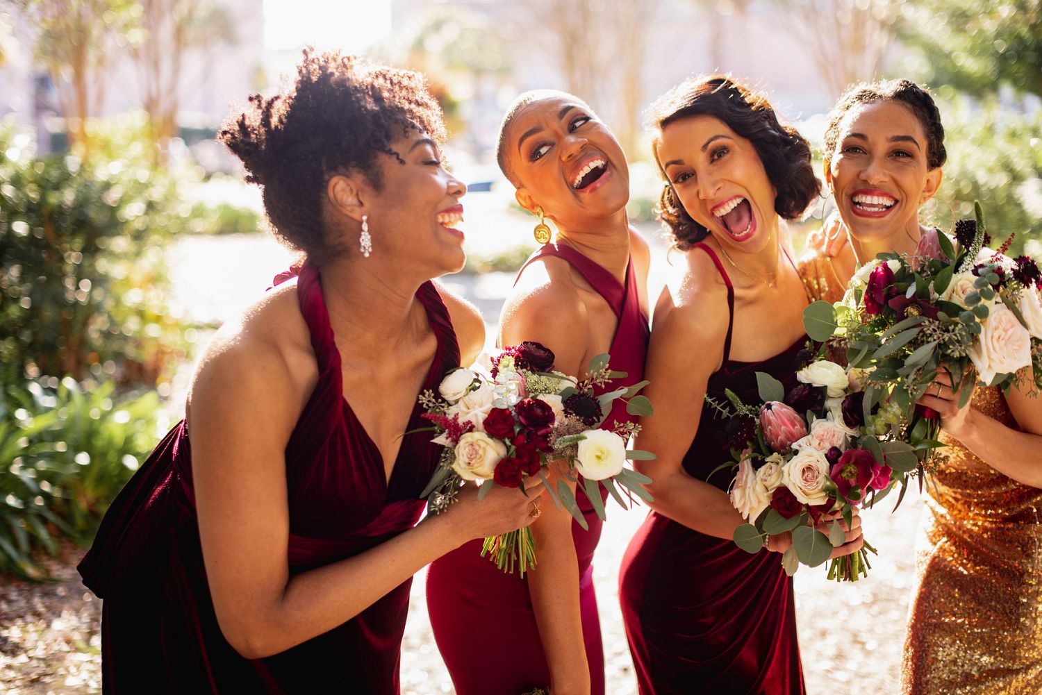 Bridesmaids in burgundy dresses laugh together while holding bouquets during outdoor wedding photoshoot.