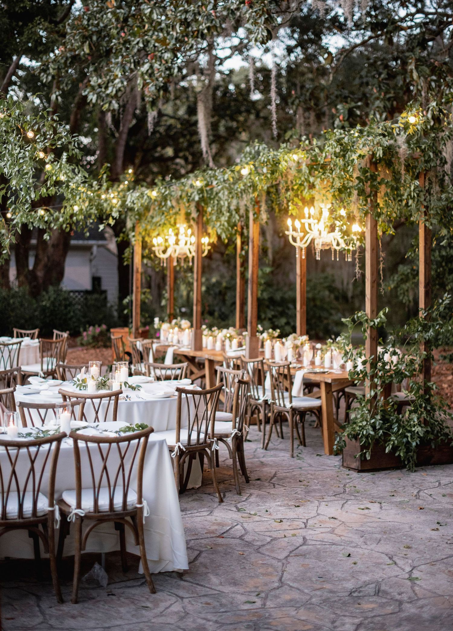 Elegant outdoor wedding reception setup with white tablecloths and wooden chairs under string lights in a garden setting.