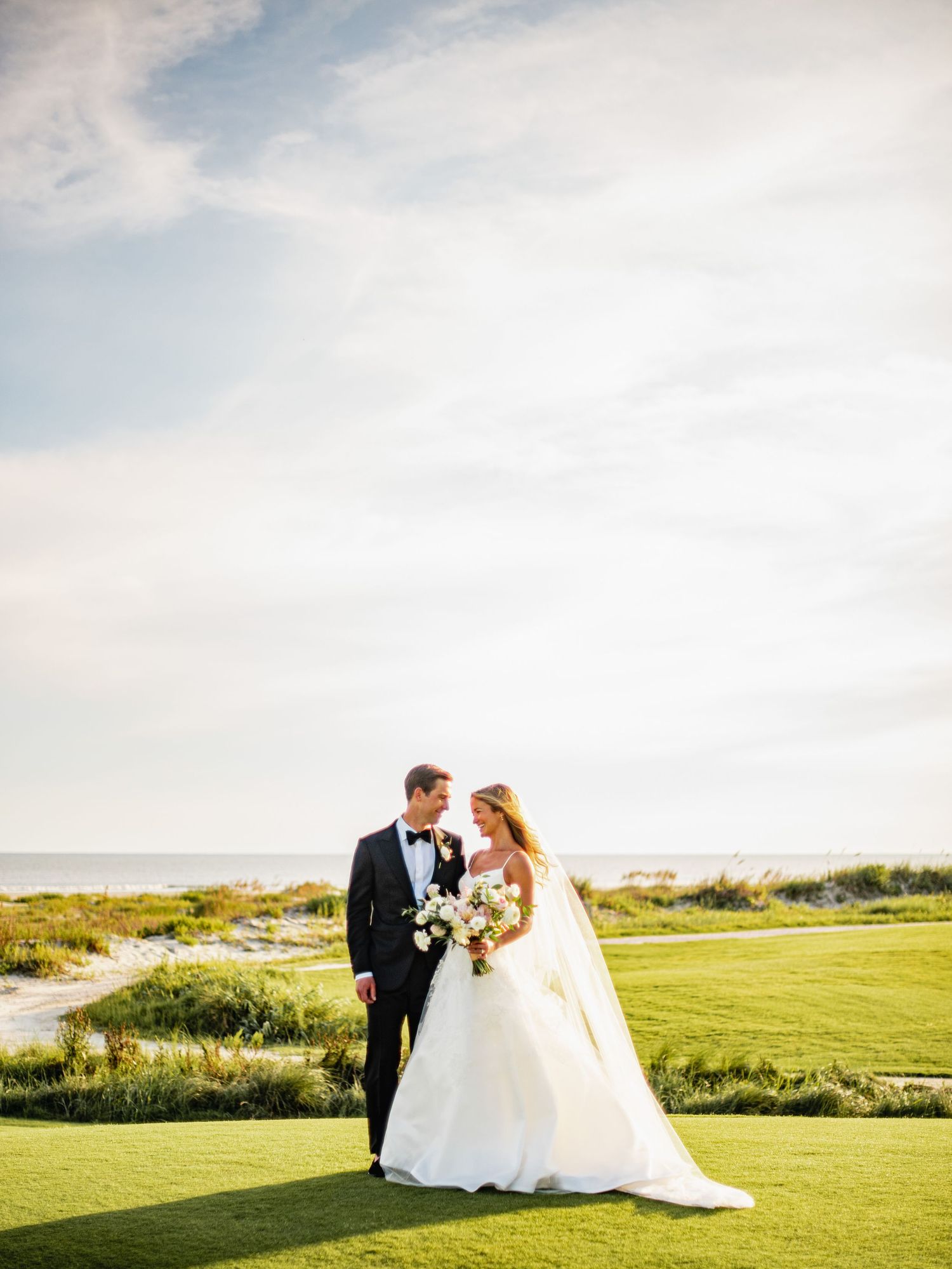 A couple in wedding attire embraces on a scenic coastal golf course with ocean views at sunset.