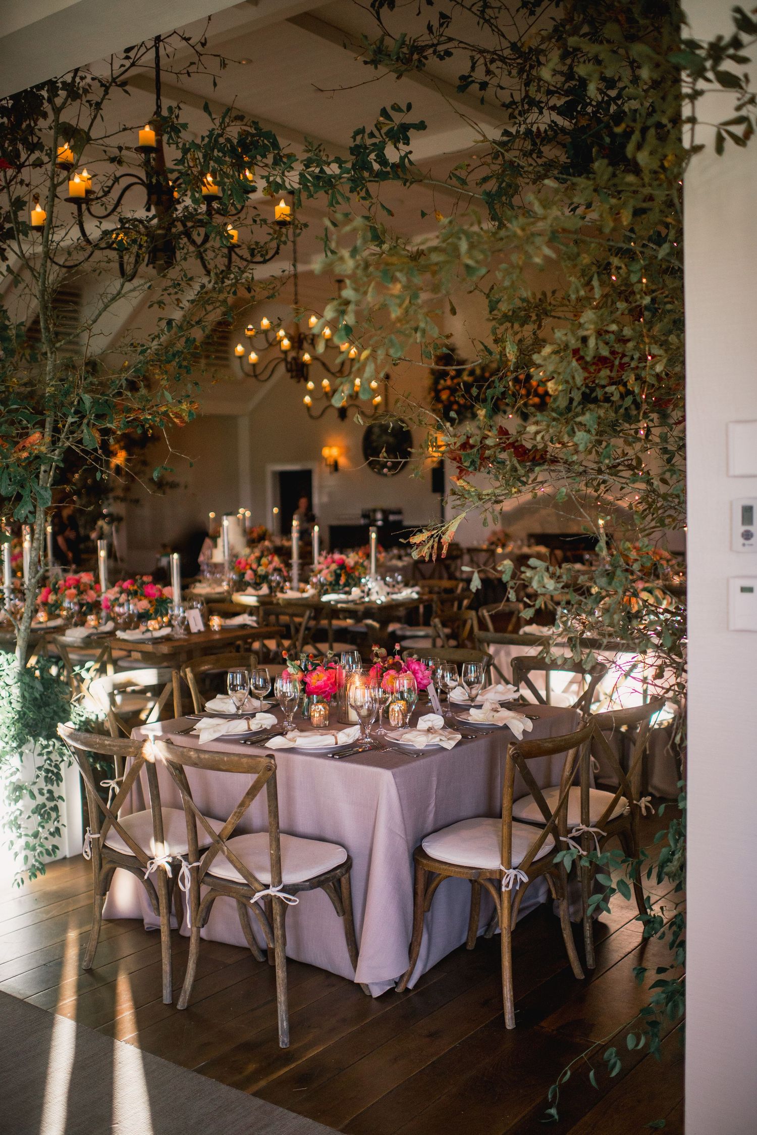 Elegant wedding reception venue with round tables, white linens, wooden cross-back chairs and hanging greenery decorations.