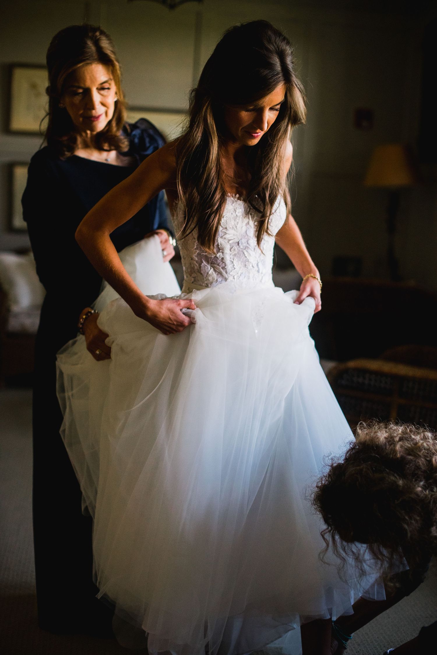 Silhouetted figures help adjust the full tulle skirt of a white wedding dress in dim lighting.