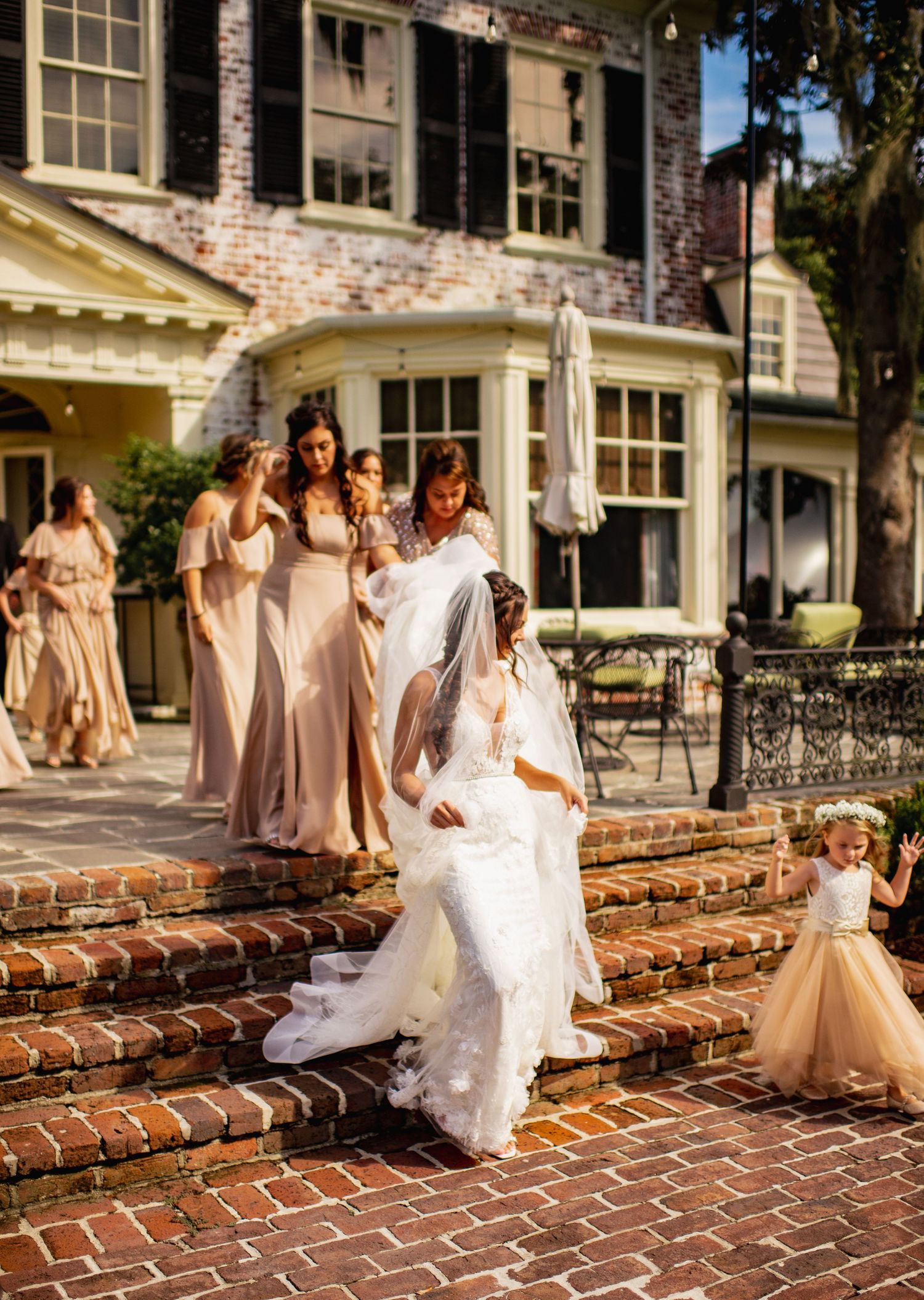 Bridesmaids in blush dresses pose with bride in white gown on brick walkway at elegant estate venue.