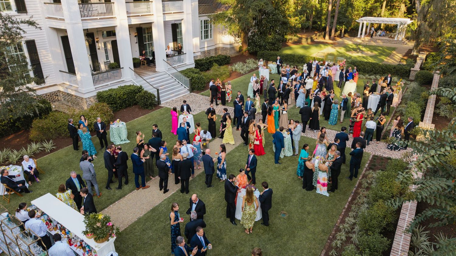 Wedding guests gather on a landscaped lawn outside a historic white building during an outdoor reception.