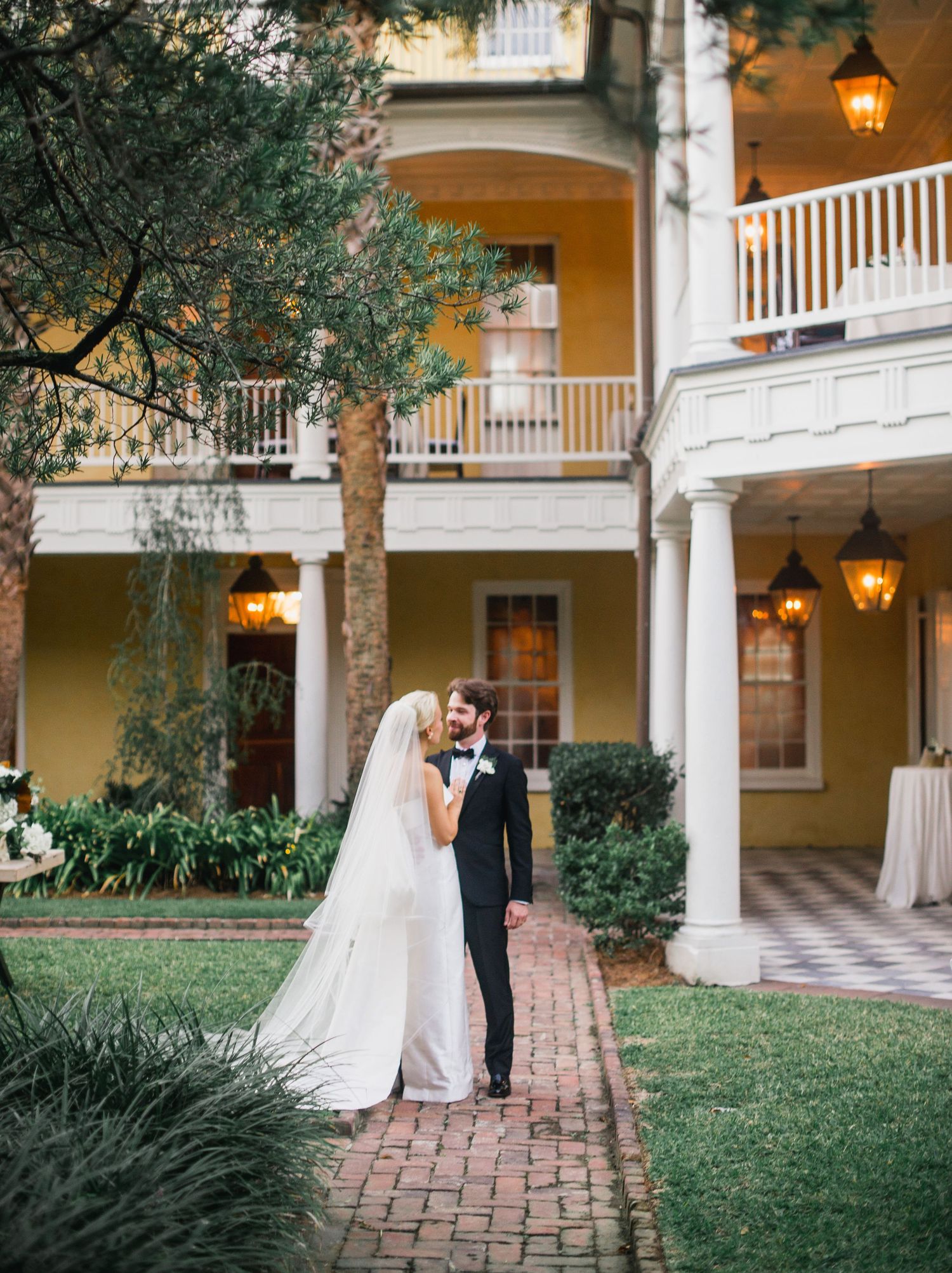 A romantic wedding couple shares intimate moments in an elegant courtyard with white columns and hanging lanterns.