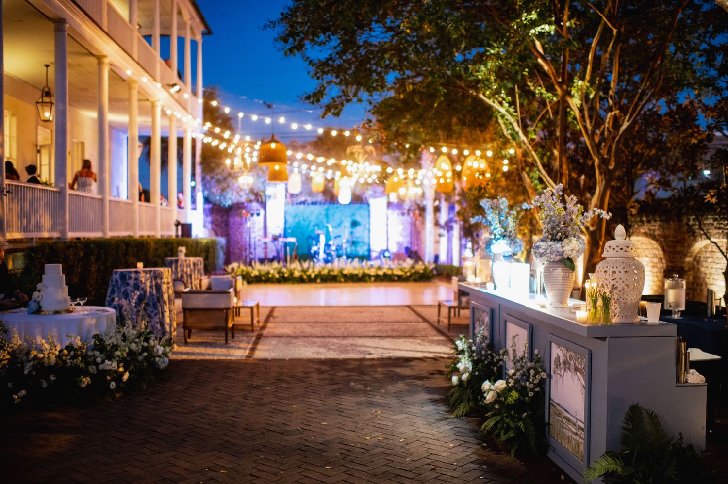 String lights illuminate an outdoor wedding reception venue at dusk with elegant white columns and glowing decorations.