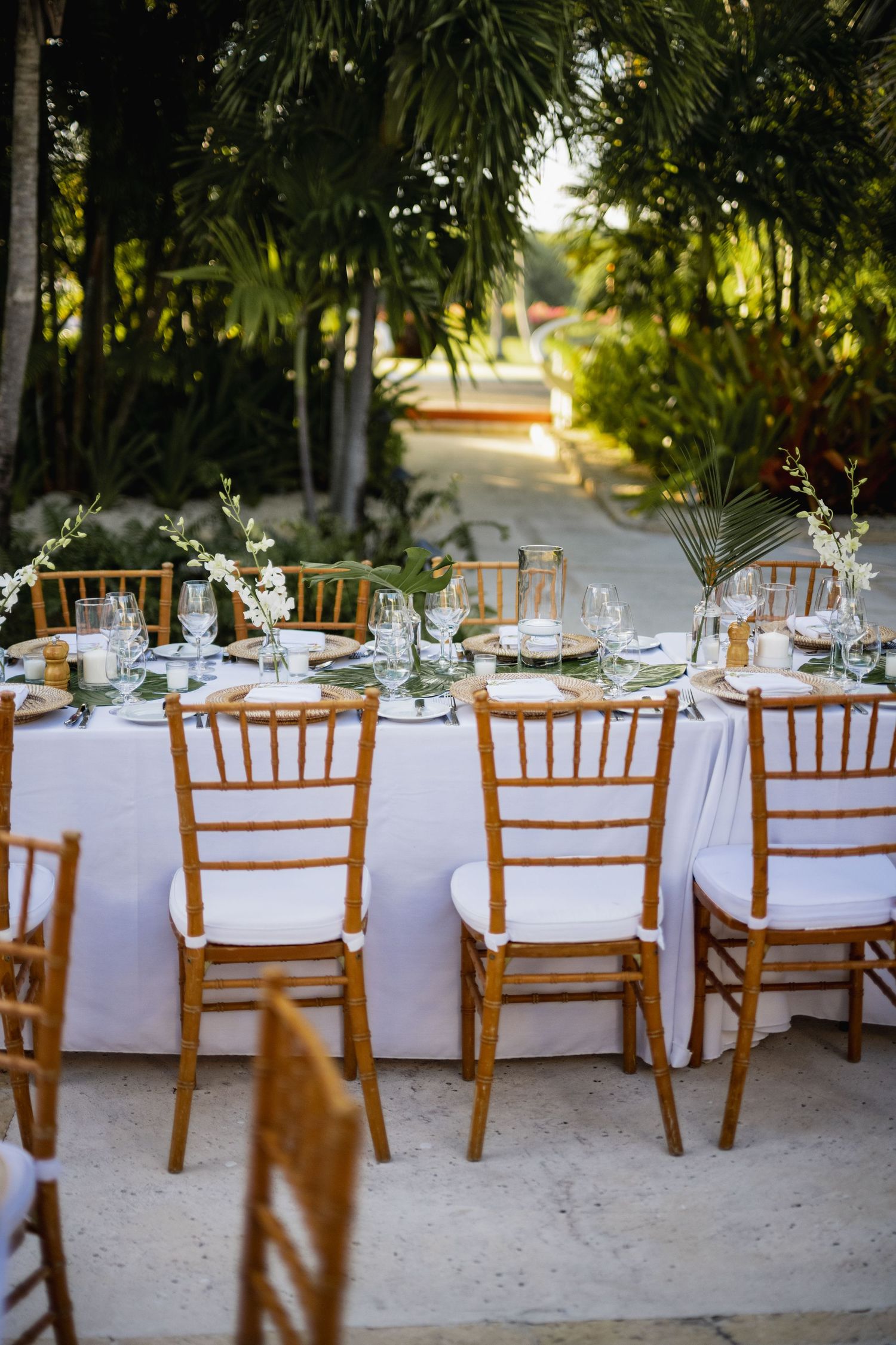 Elegant outdoor wedding reception setup with wooden chiavari chairs and white linens under tropical palm trees.