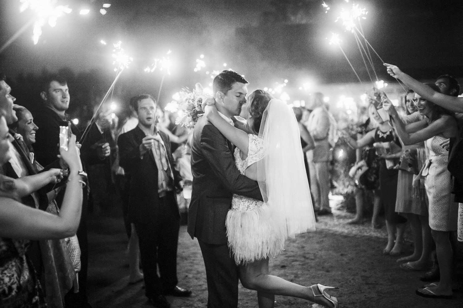 Wedding guests hold sparklers creating a magical illuminated pathway at an outdoor nighttime celebration.