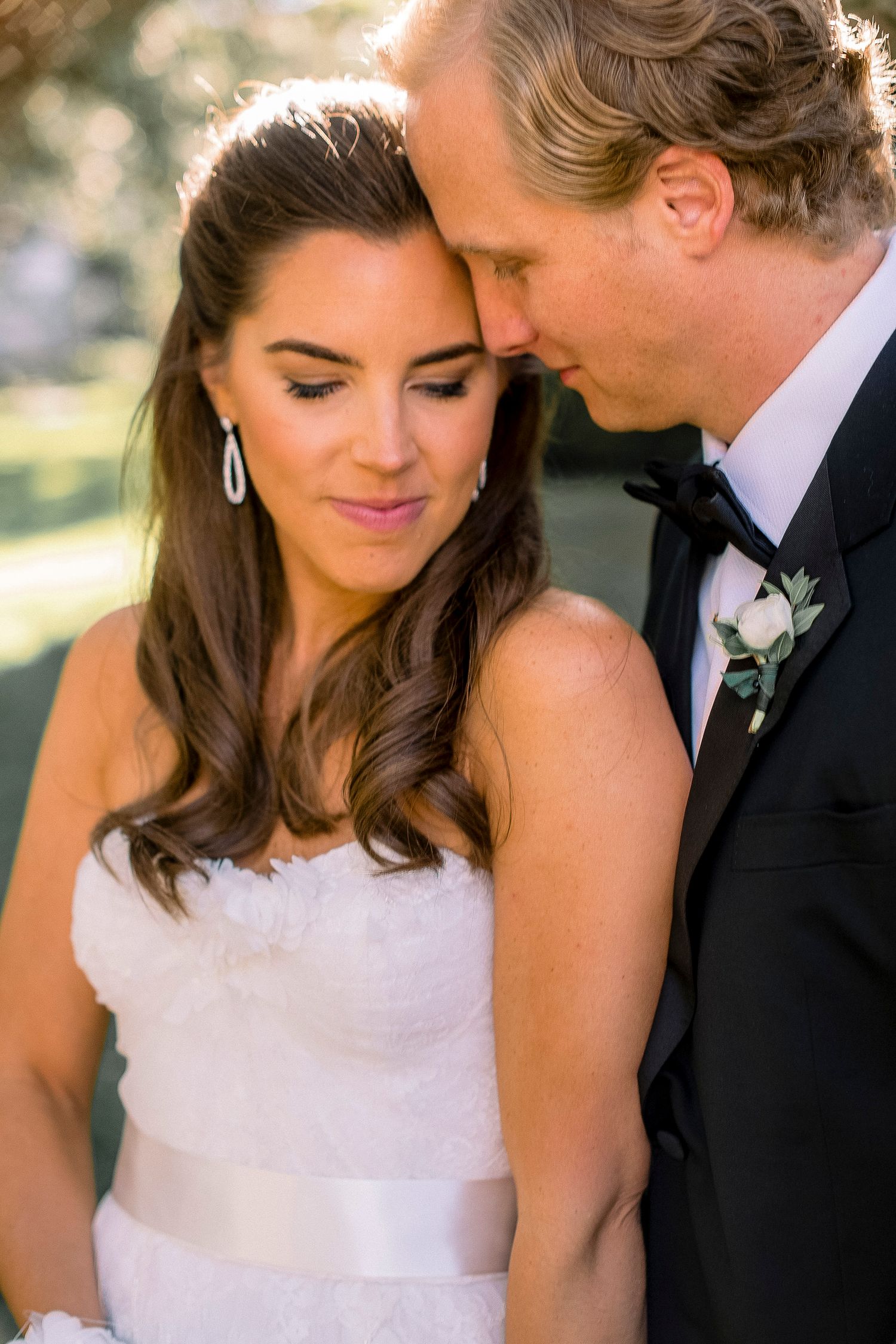 Intimate wedding portrait of couple in formal attire against sunlit background.