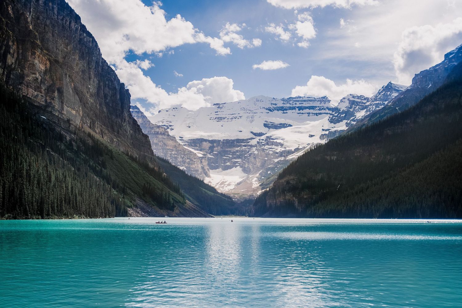 Snow-capped mountain peaks reflect in the turquoise waters of Lake Louise surrounded by evergreen forest.