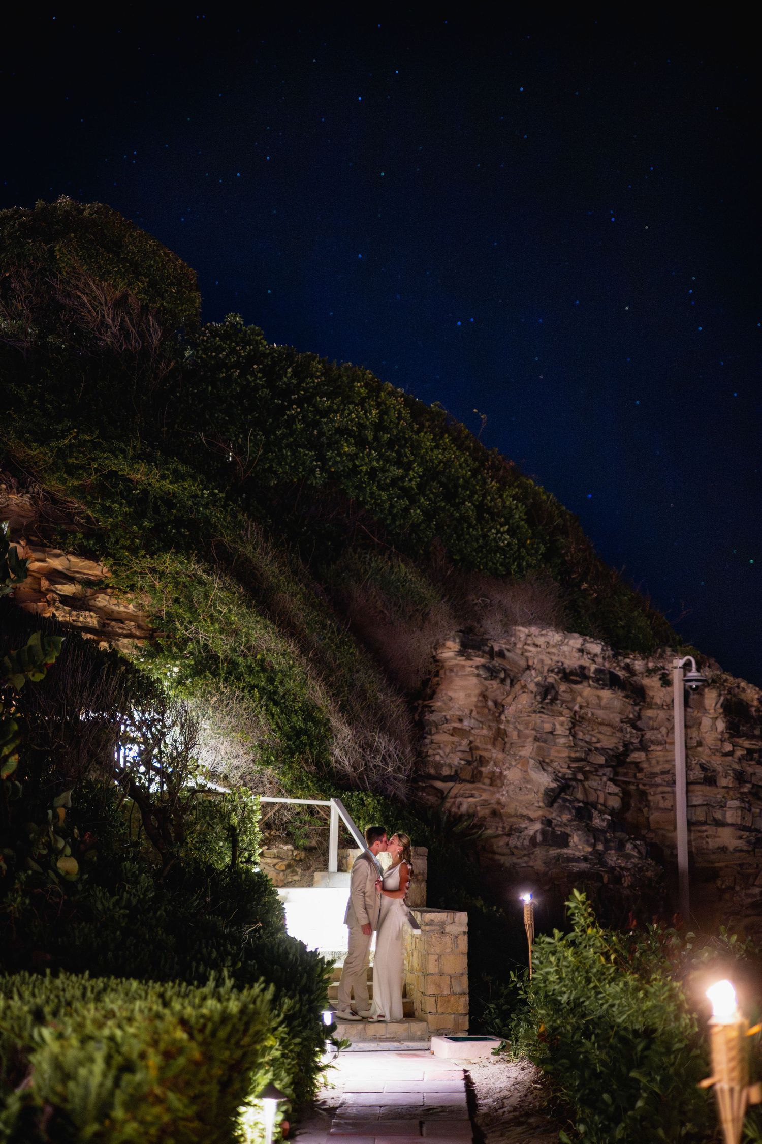 Stone pathway lit by torches and lights leading through a tropical garden at night.