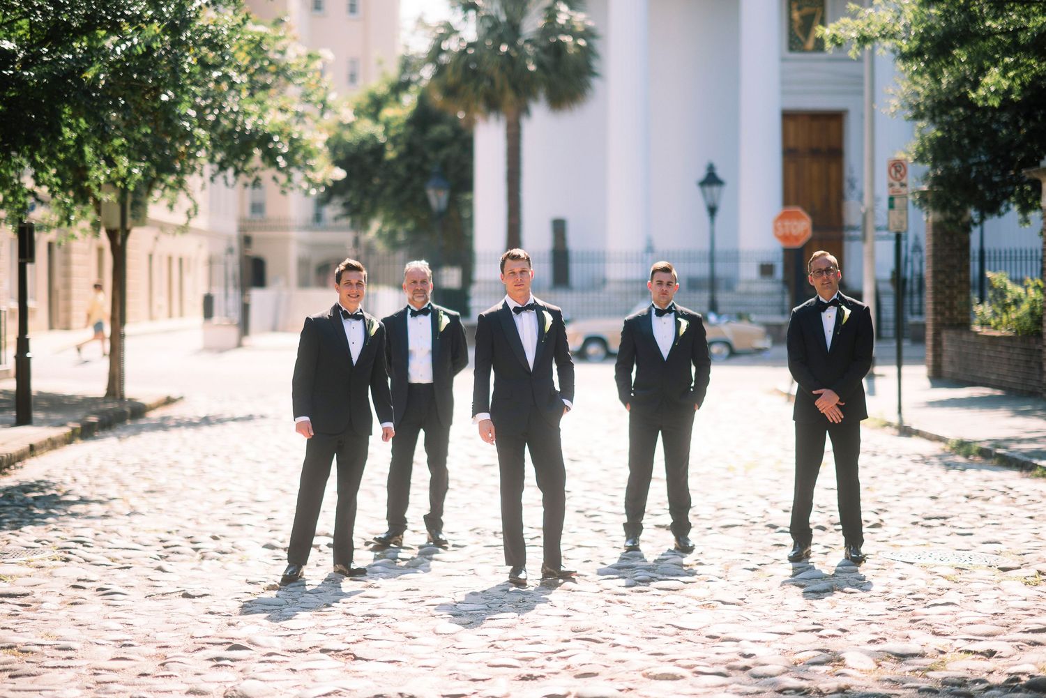 Wedding party in black tuxedos walking along a sunny outdoor pathway lined with trees and buildings.