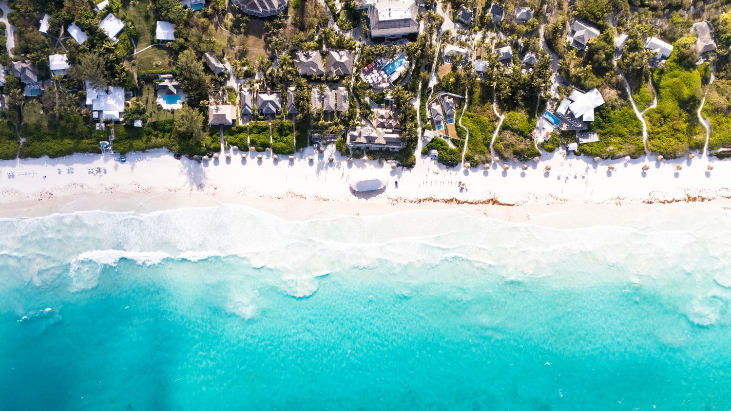 Aerial view of turquoise ocean waves crashing on white sandy beach with lush tropical vegetation along coastline.