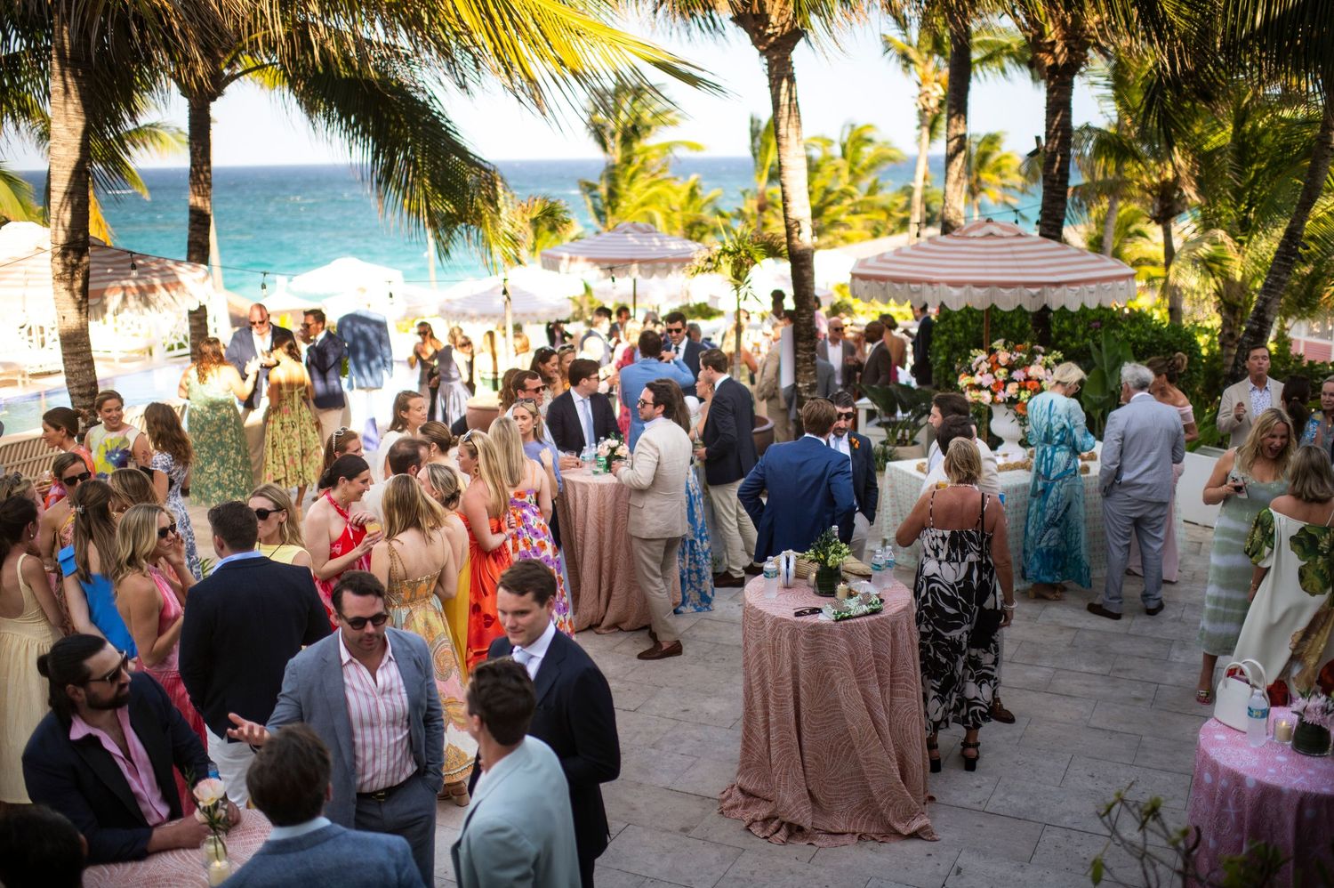 Guests mingle at an elegant outdoor beach wedding reception under palm trees at sunset.