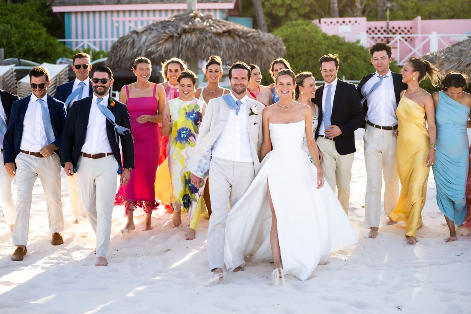 Wedding party members in white and pastel attire walk together in a line outdoors during a celebration.