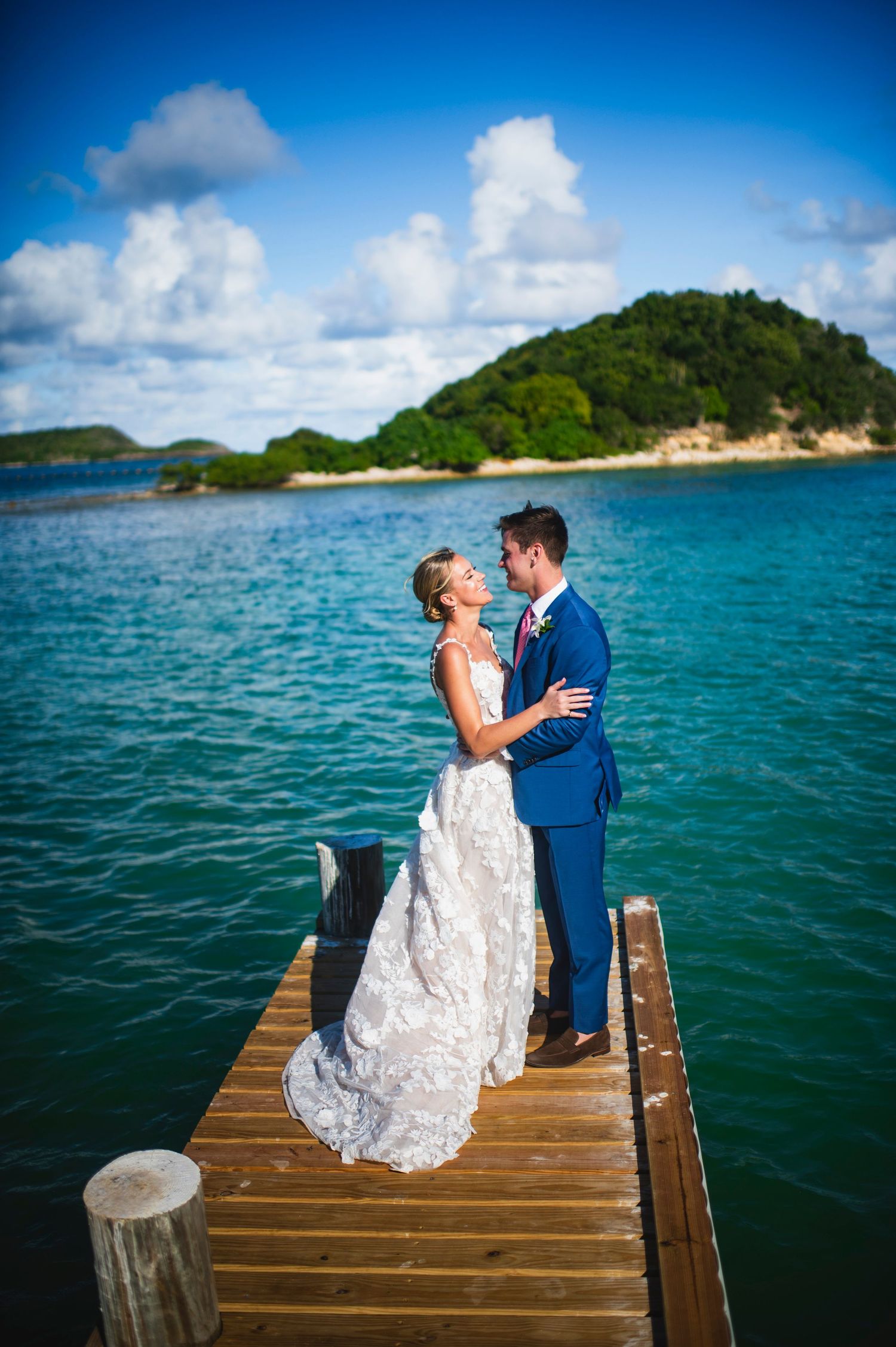 A couple in wedding attire shares a romantic moment on a wooden dock overlooking turquoise tropical waters and green islands.