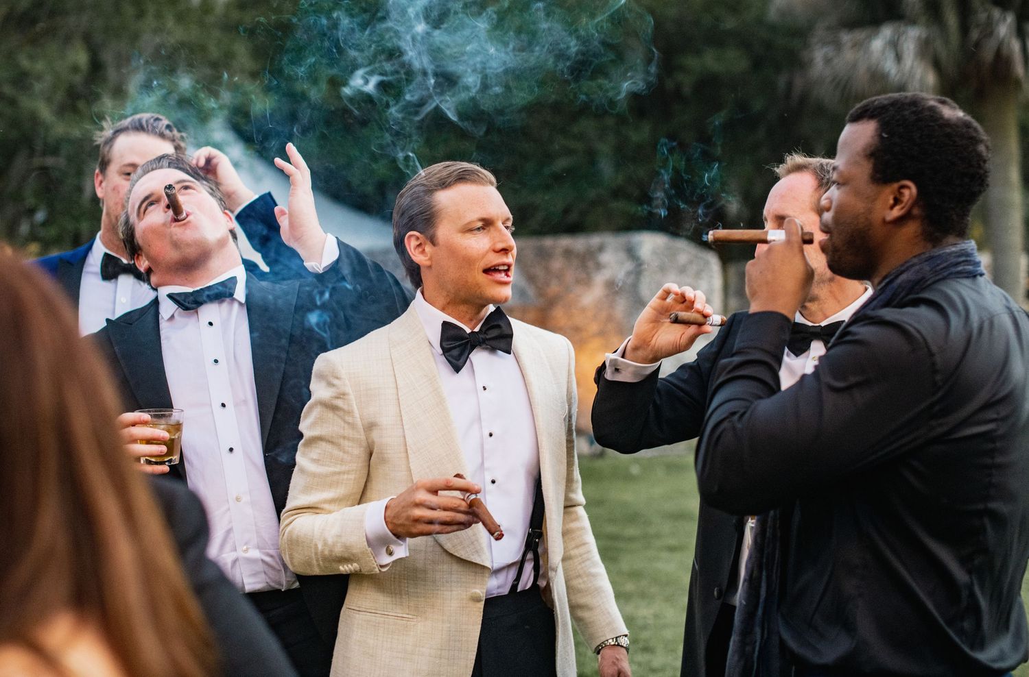 Group enjoying cigars and drinks at an outdoor celebration with smoke wafting through the air.