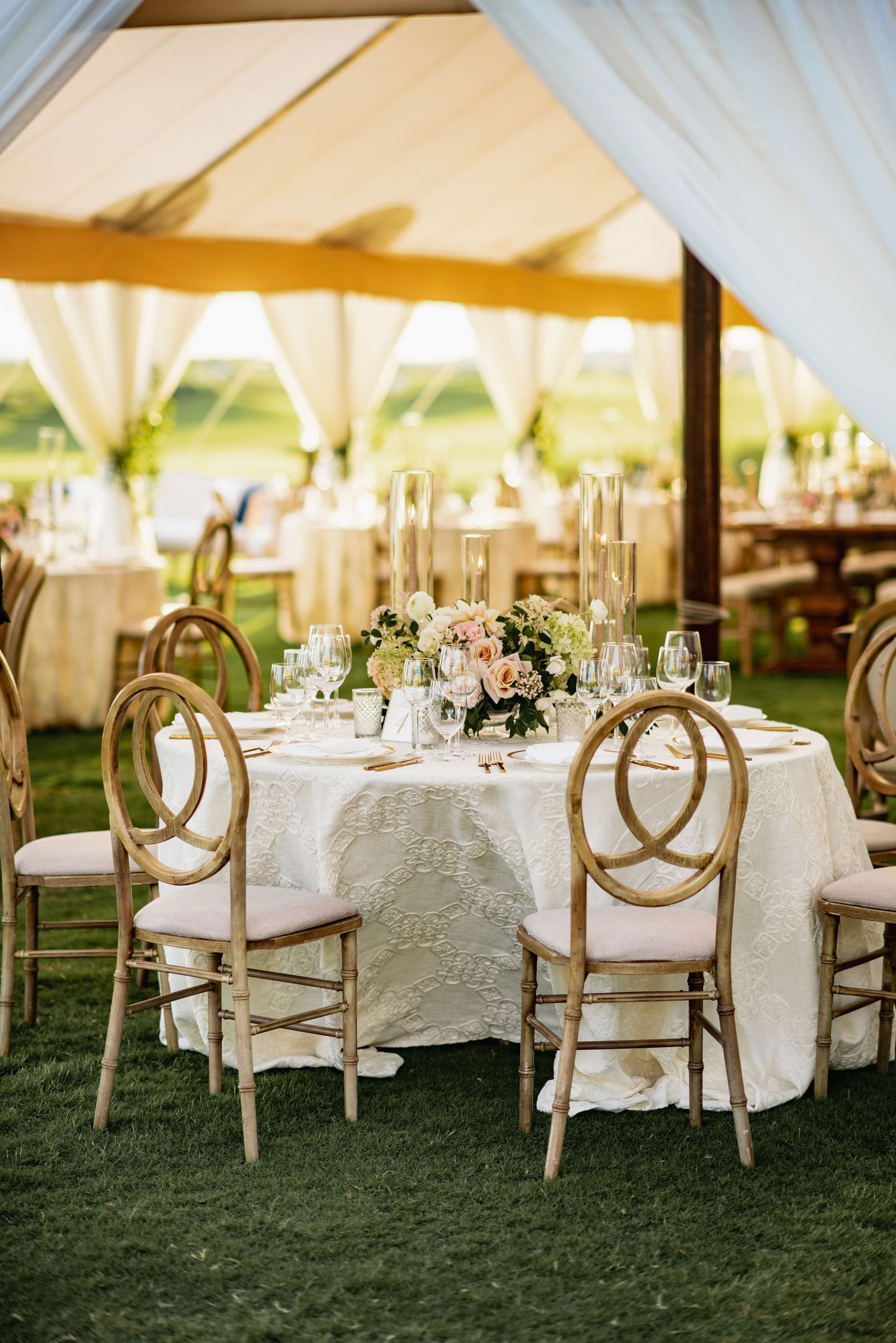 Elegant outdoor wedding reception setup with white linens, floral centerpieces and round-back chairs under a white tent.