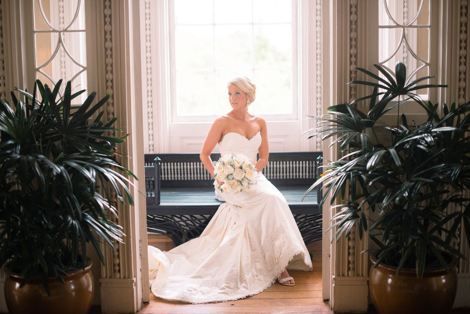 A bride in a white strapless wedding gown holds a bouquet while standing between potted palm plants near a window.